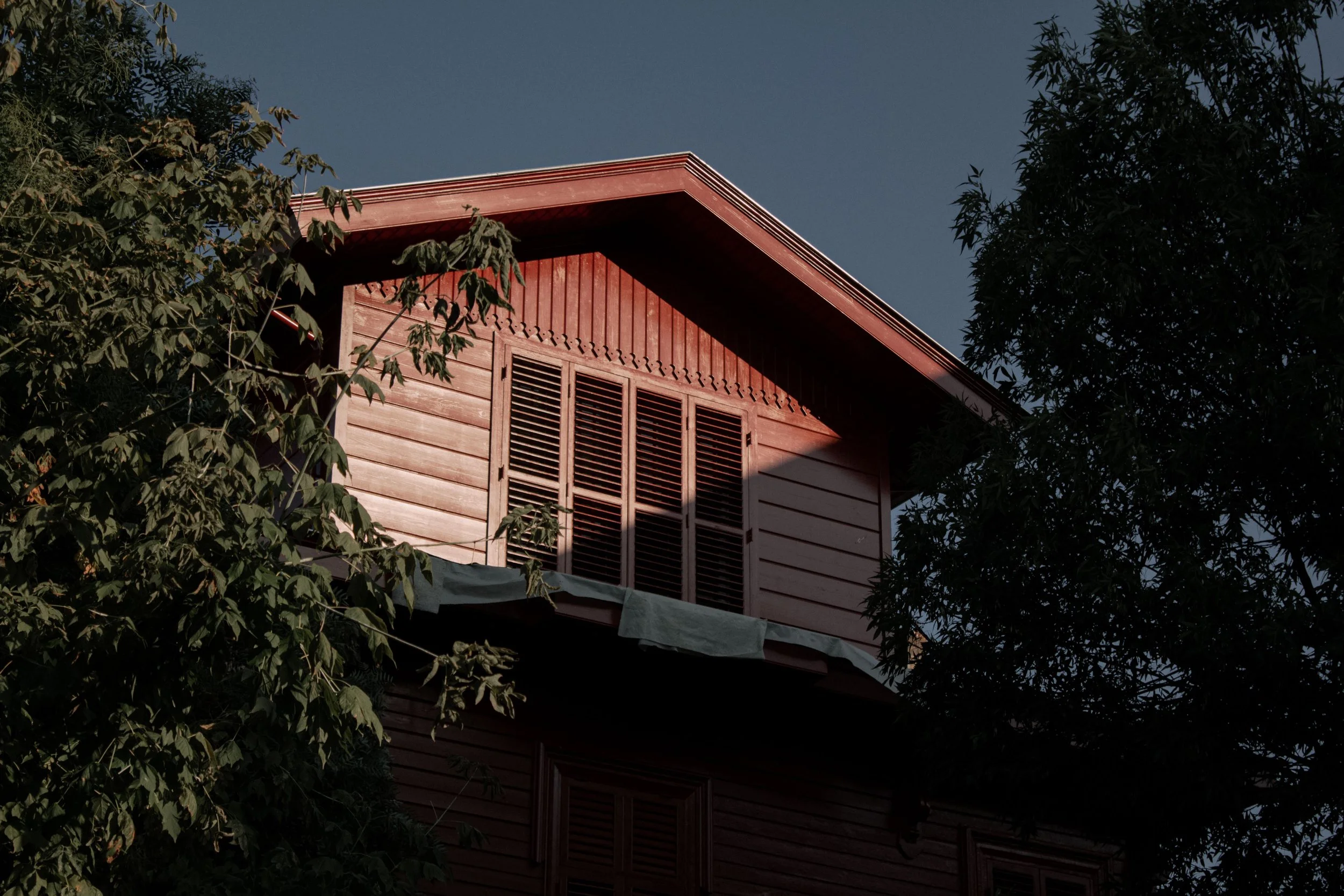 A red wooden house with closed shutters, surrounded by green trees and partially shaded, under a clear sky.
