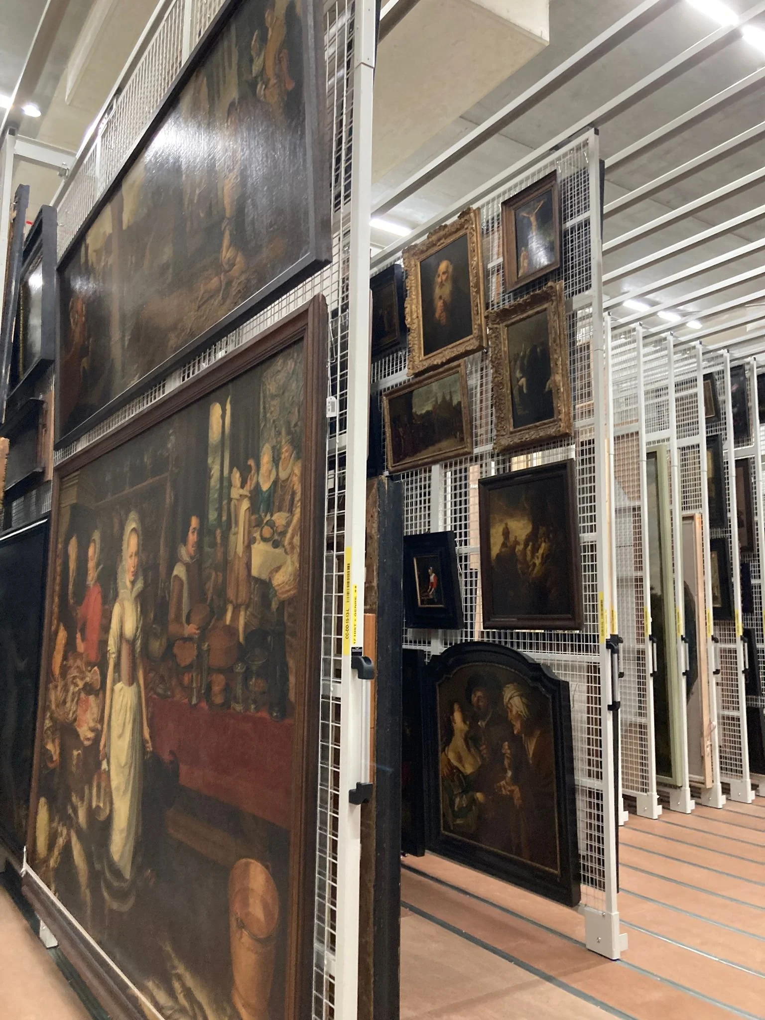 View of art paintings on display on a metal grid shelving unit in a store or gallery, including a large painting on the left and several smaller framed paintings on the right.