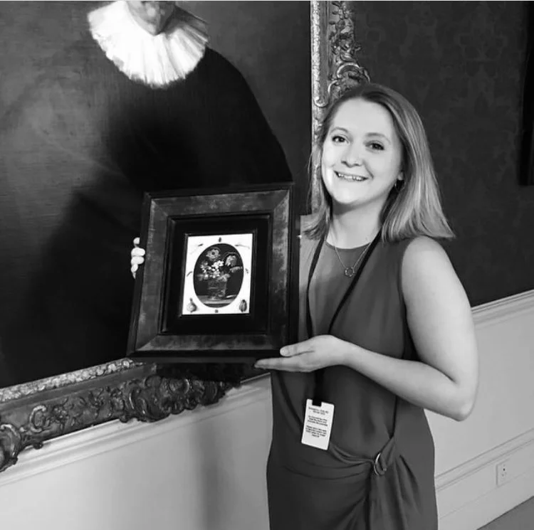 A young woman with shoulder-length hair is smiling and holding a framed artwork in front of a large portrait painting inside a gallery or museum.