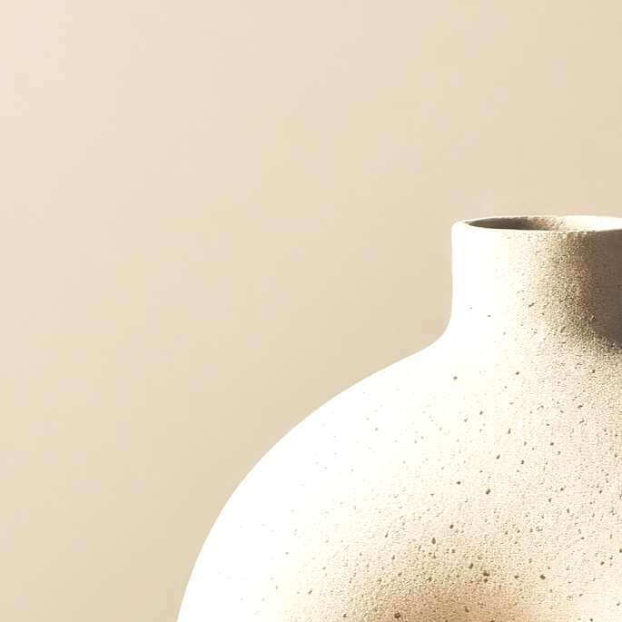 A close-up of a textured, off-white ceramic vase with a rounded body and a narrow neck against a plain, beige background.