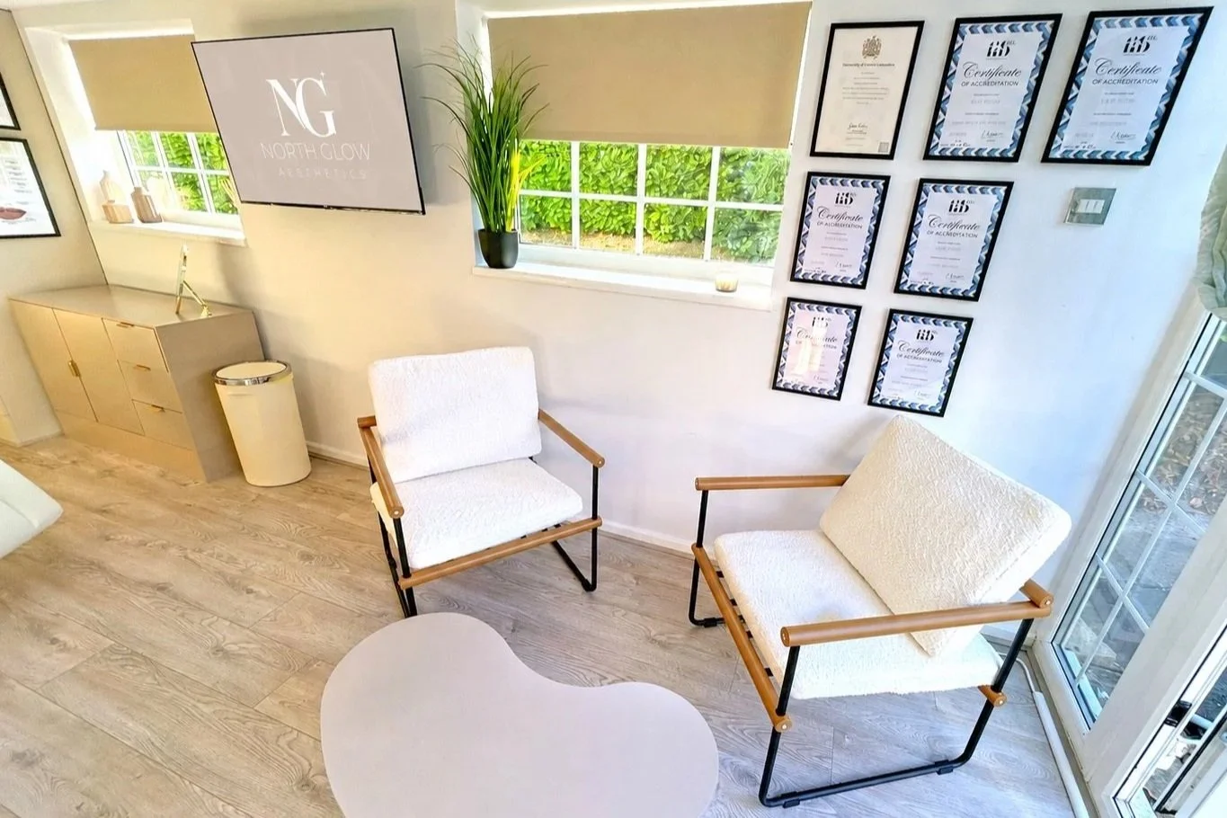 Consultation area at North Glow Aesthetics with cream boucle armchairs, a coffee table, and Katie's accreditation certificates displayed on the wall, Norden, Rochdale.