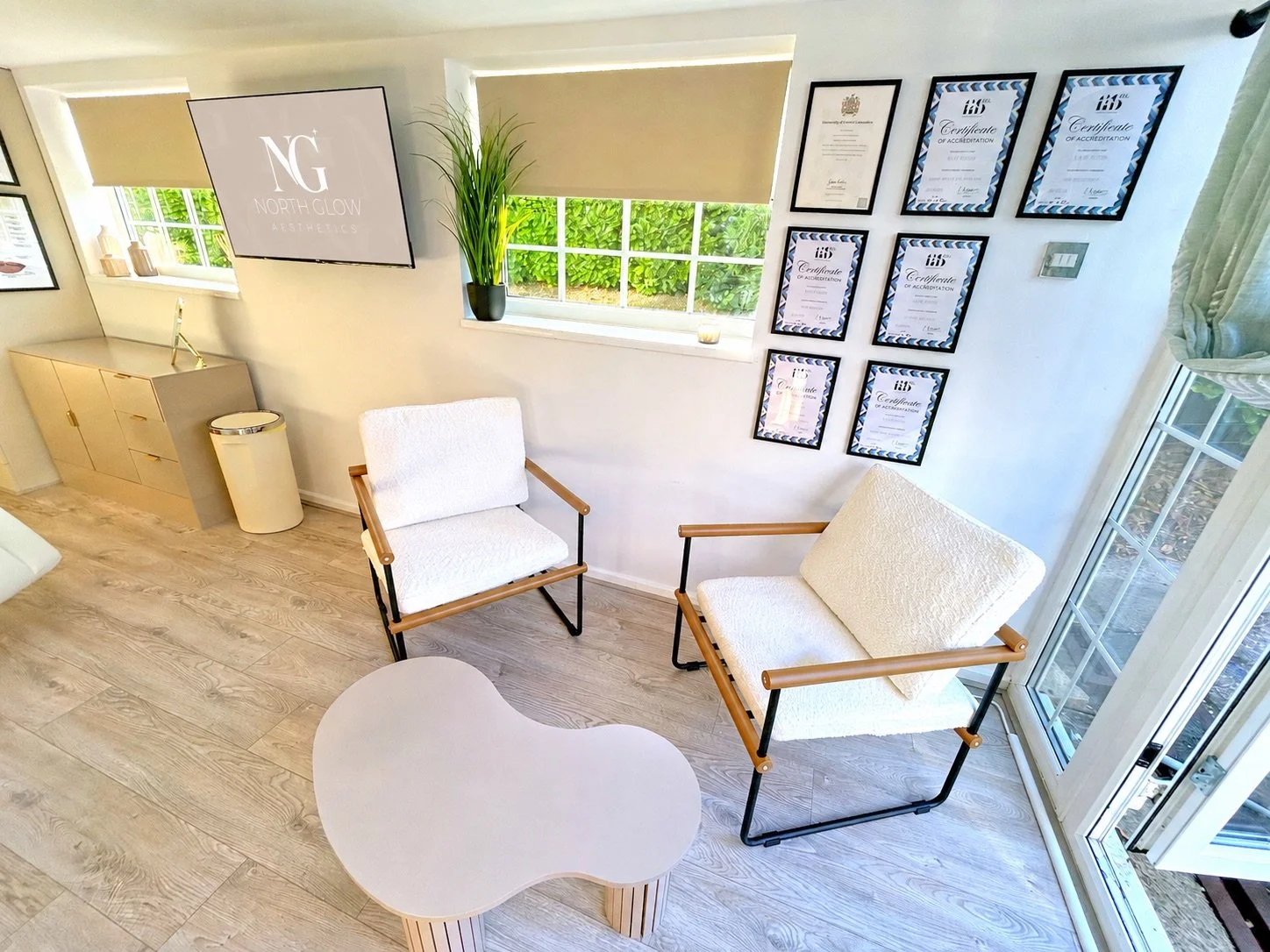 Consultation area at North Glow Aesthetics with cream boucle armchairs, a coffee table, and Katie's accreditation certificates displayed on the wall, Norden, Rochdale.