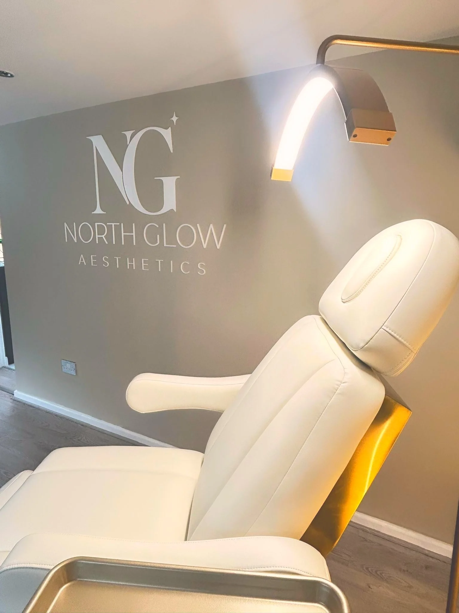 Close-up of the illuminated gold arc lamp above the treatment chair headrest at North Glow Aesthetics, with the NG wall logo beside it, Norden, Rochdale.