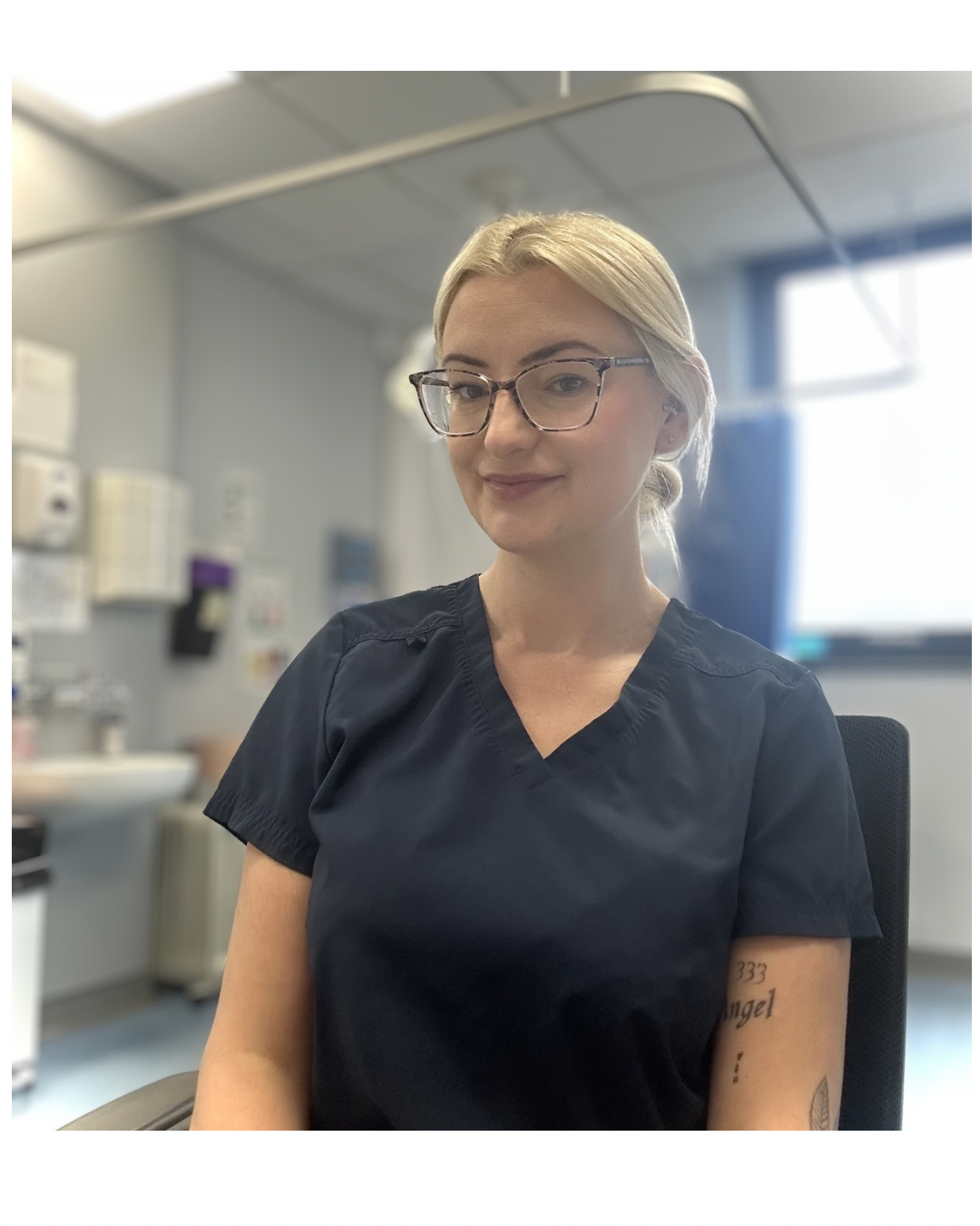 A woman with blonde hair, glasses, and tattoos wearing medical scrubs in a clinical setting with medical equipment and a monitor in the background.