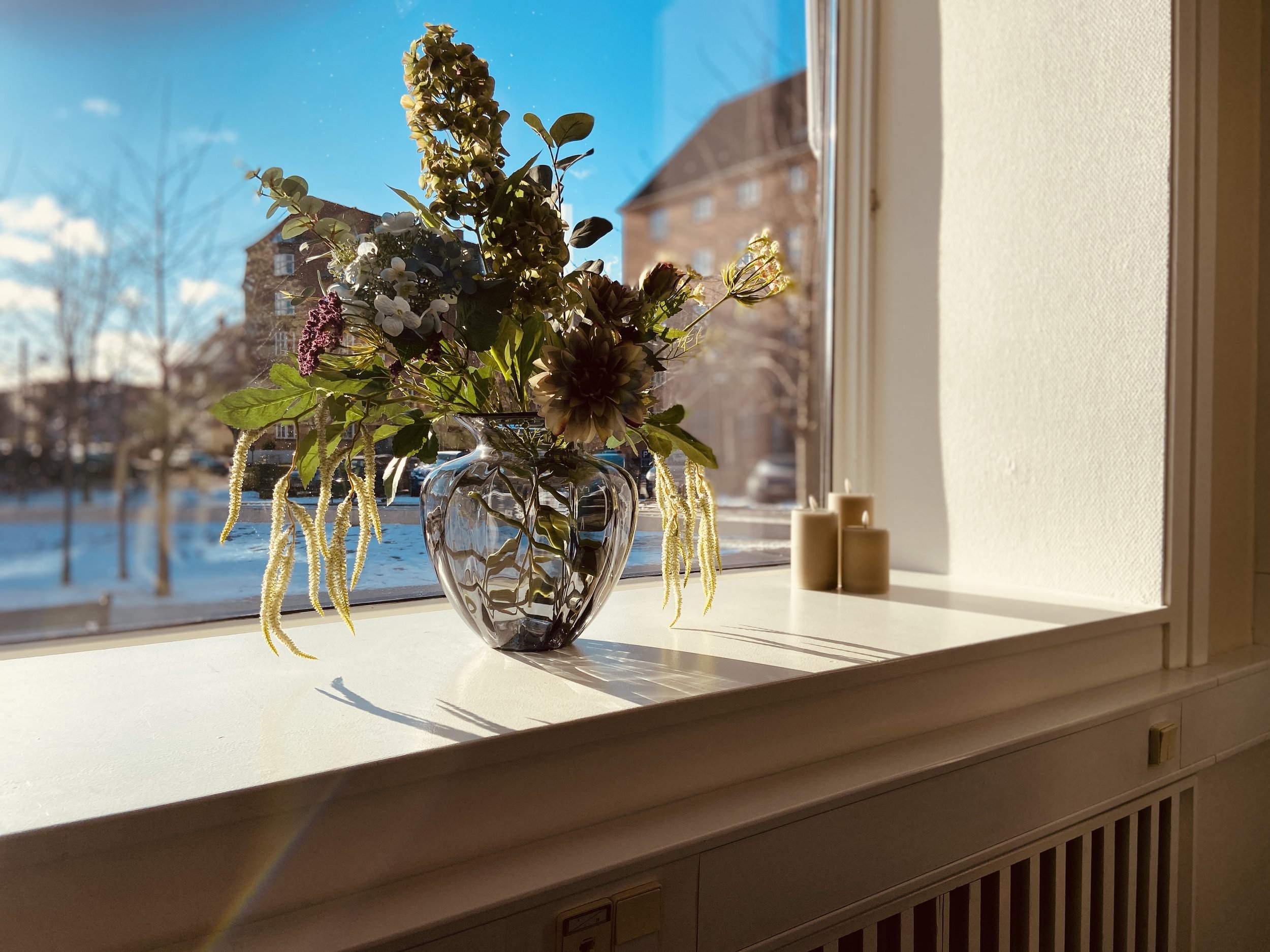 En vase med blomster på en vindueskarm med udsigt til en klar blå himmel og bygninger i baggrunden.