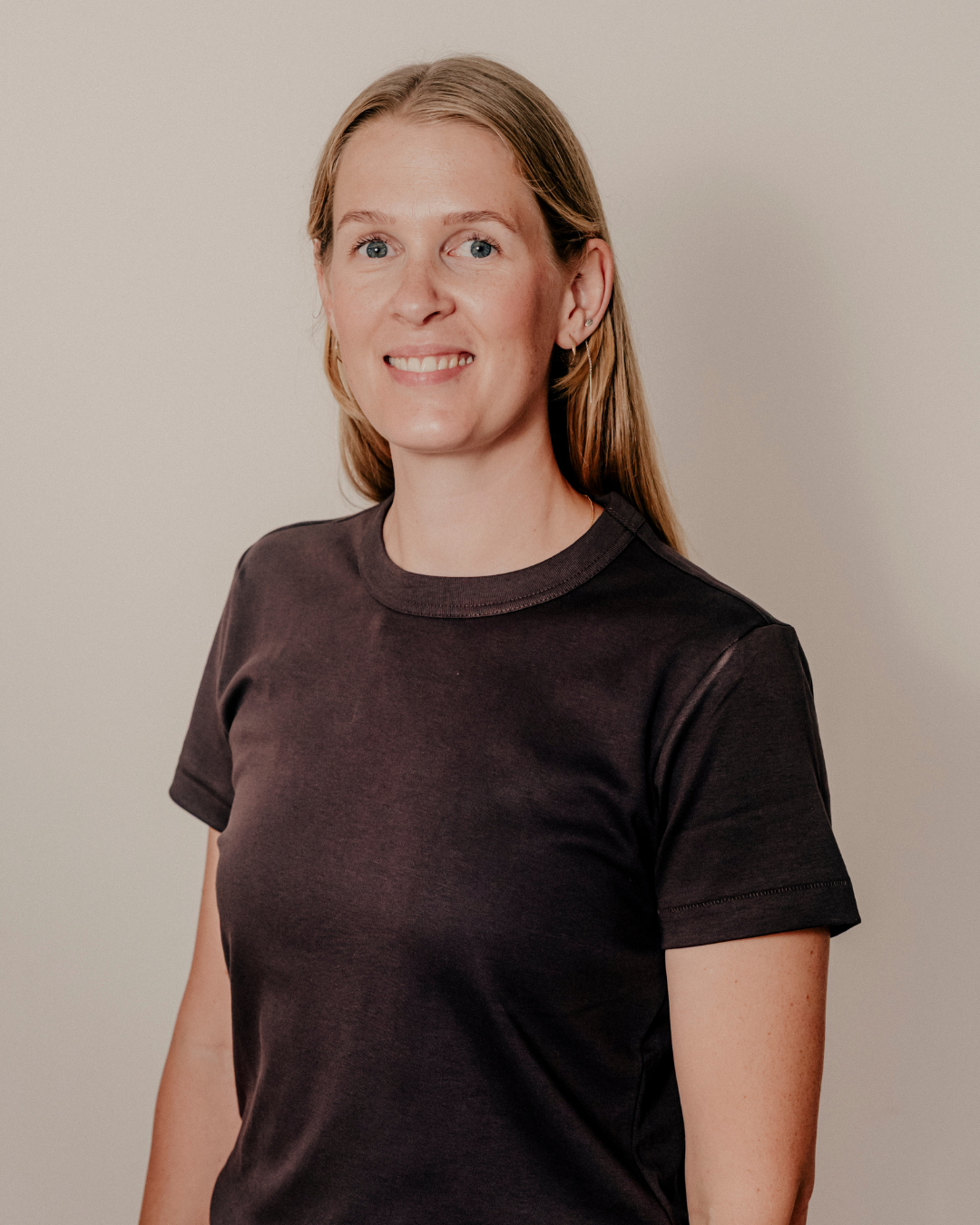 En ung kvinde med lyst hår iført en sort T-shirt, står foran en neutral baggrund, smilende mod kameraet.