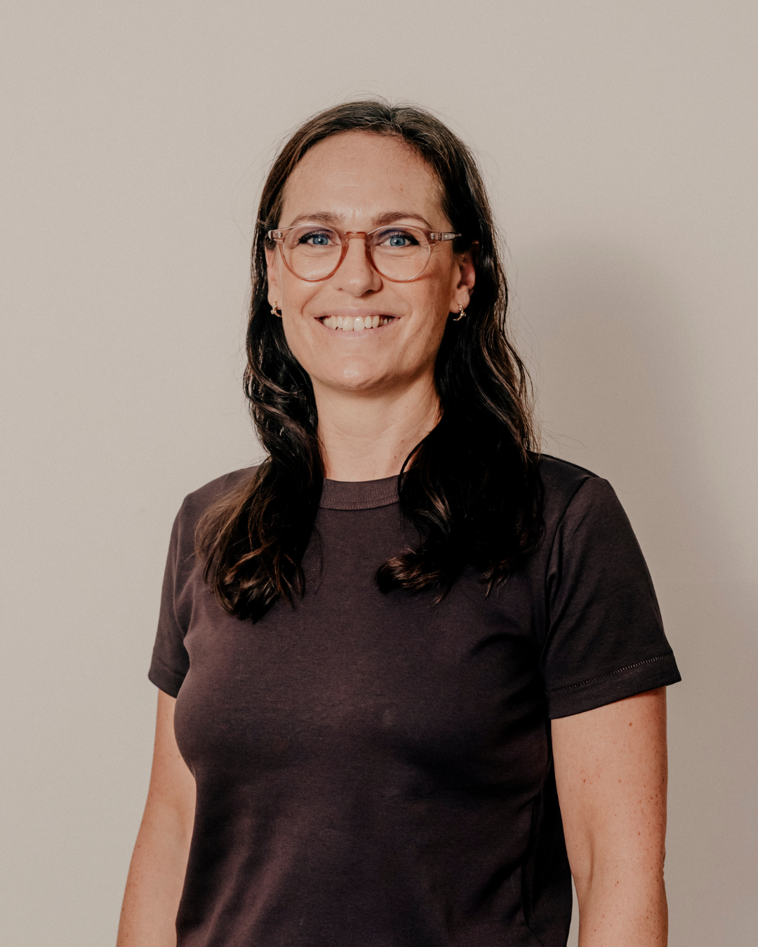 Portræt af en smilende kvinde med brunt hår, briller og en sort T-shirt, står foran en simpel, lys baggrund.