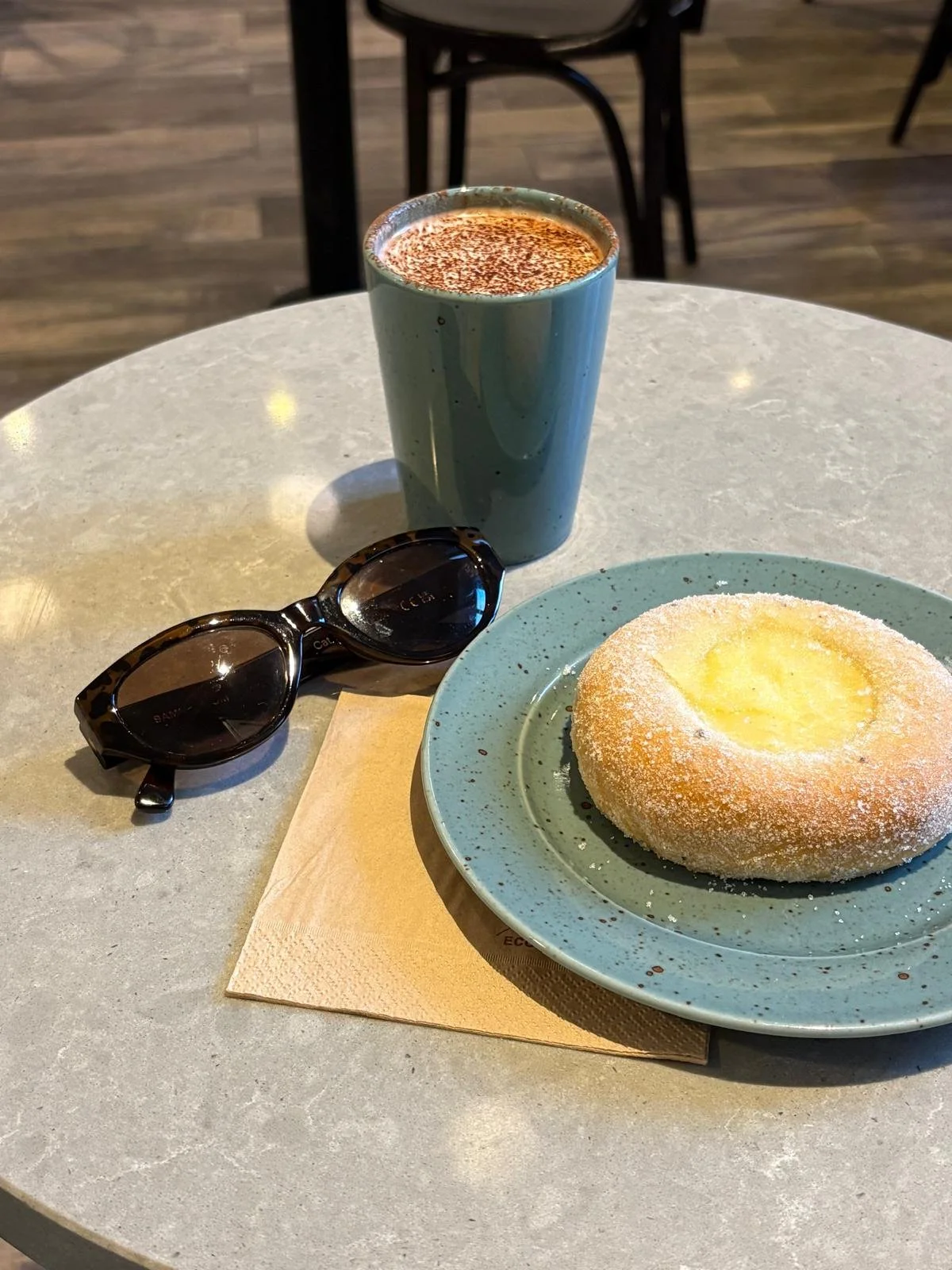 Een tafeltje in een café met een kop chocomelk, een gebakje en een zonnebril.