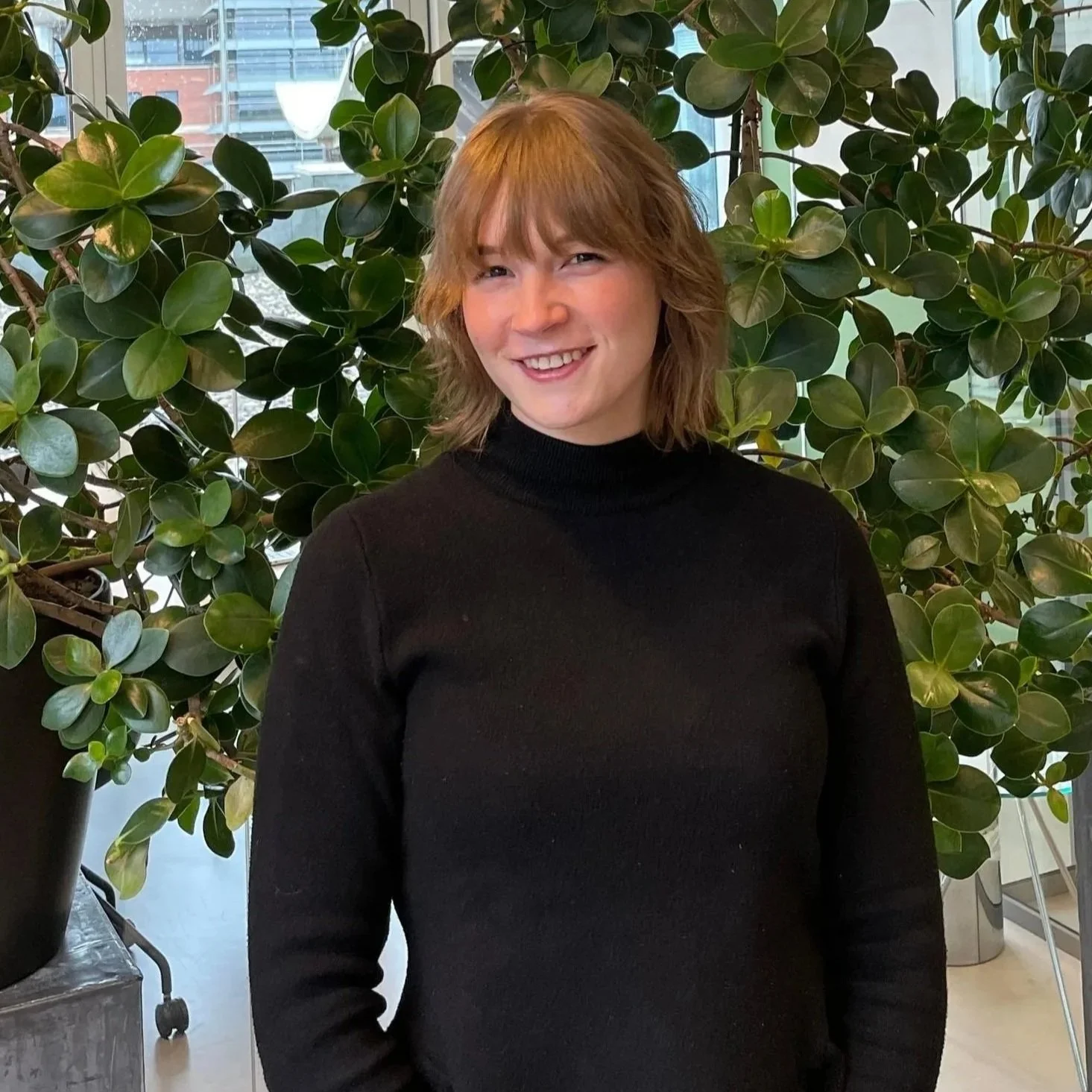 Freelance journalist Lobke Vermeer voor een achtergrond van groene planten staat met een zwart shirt aan en een lach op haar gezicht.
