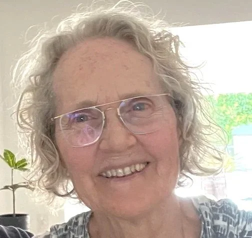 Robyn Seamer A smilingwoman with curly gray hair, wearing glasses and a patterned shirt, standing indoors near a window with a green plant visible in the background.