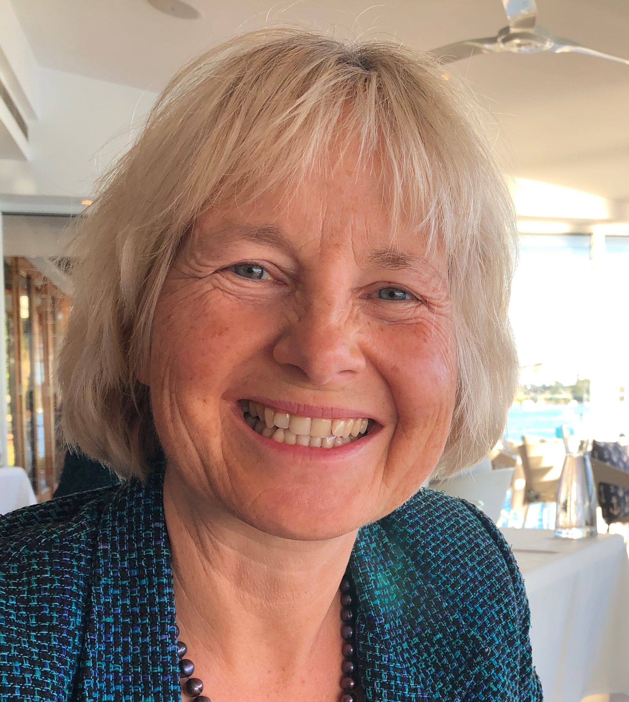 Close-up of Manuela Da Rin with light blonde hair, smiling, showing white teeth, wearing a blue patterned jacket and a black beaded necklace, with a background of large windows and outdoor scenery.