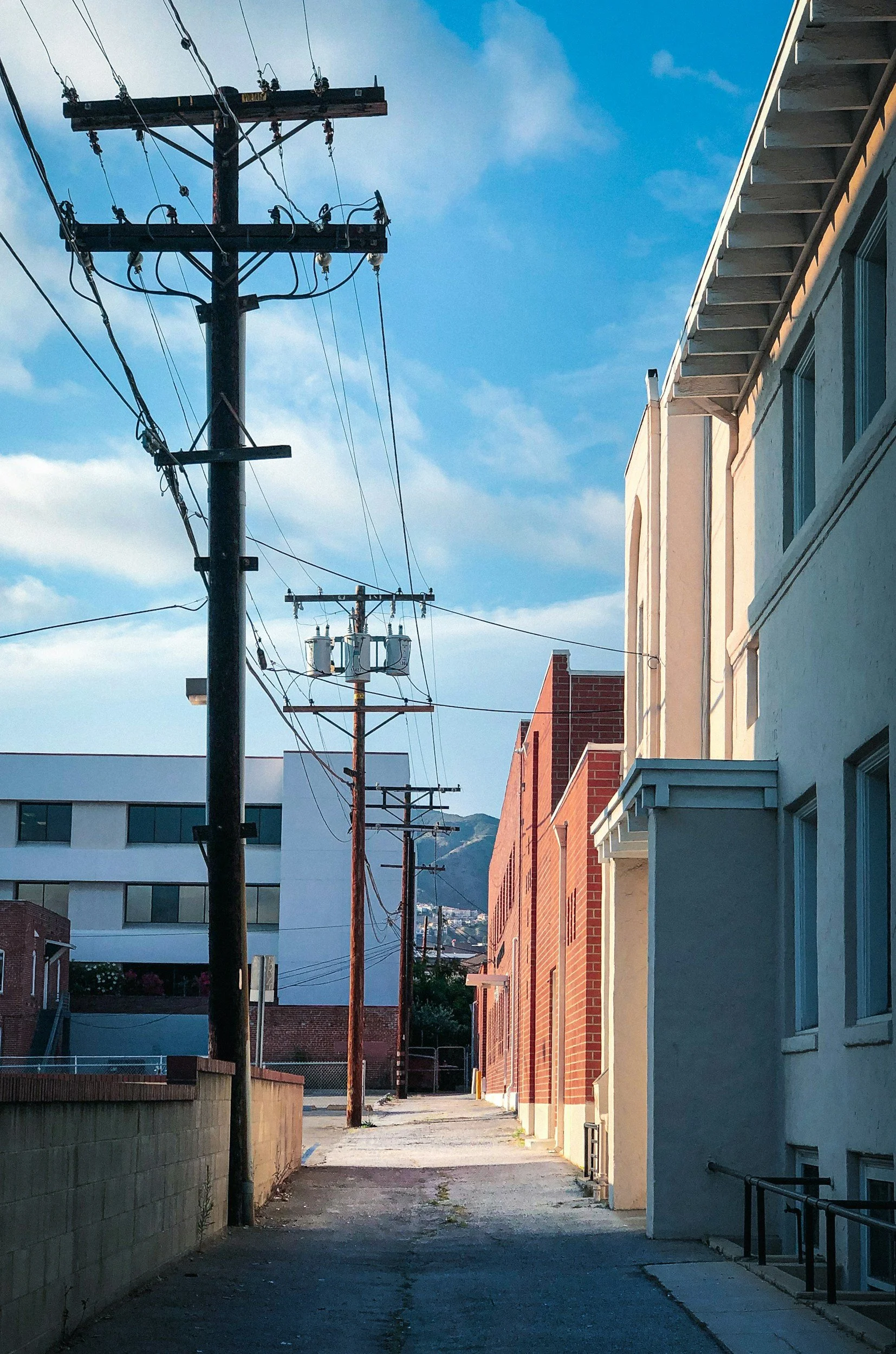 An alleyway between buildings with power lines overhead, some buildings are made of brick and others are painted in light colors. The sky is partly cloudy.