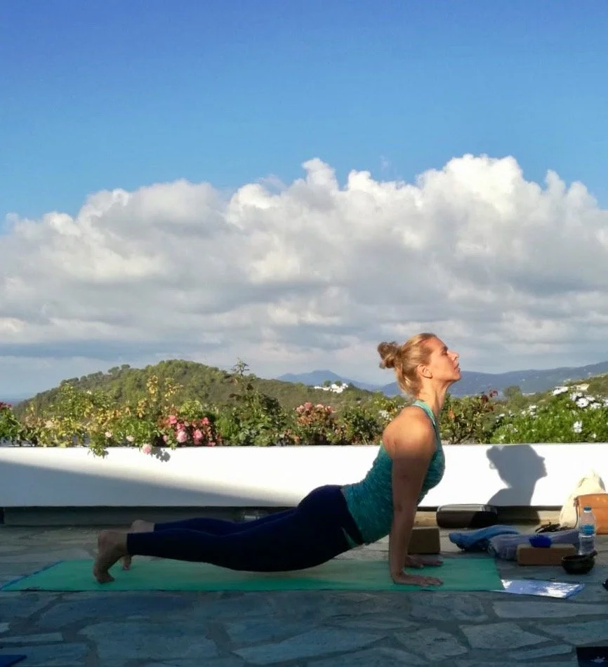 Vrouw doet yoga op een mat buiten met uitzicht op bergen en een plek met bloemen, onder een bewolkte blauwe lucht.