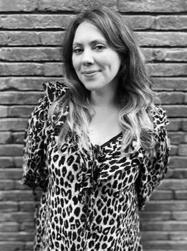 A woman with wavy hair wearing a leopard print top standing in front of a brick wall.