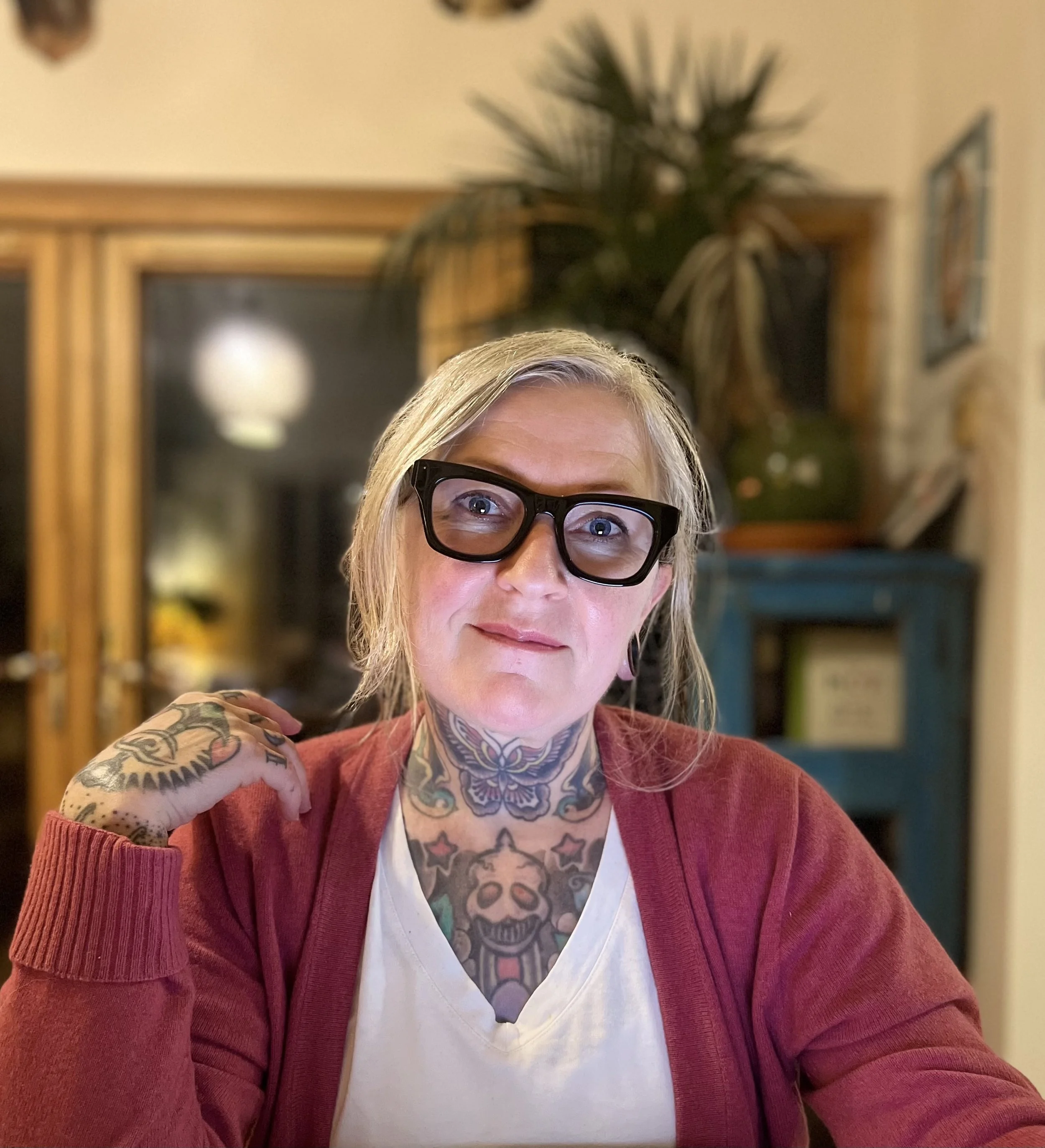 Close-up of an older woman wearing large black glasses, a white shirt, and a red cardigan, showing colorful tattoos on her chest and arm. She is in a room with a wooden cabinet, a plant, and framed pictures in the background.