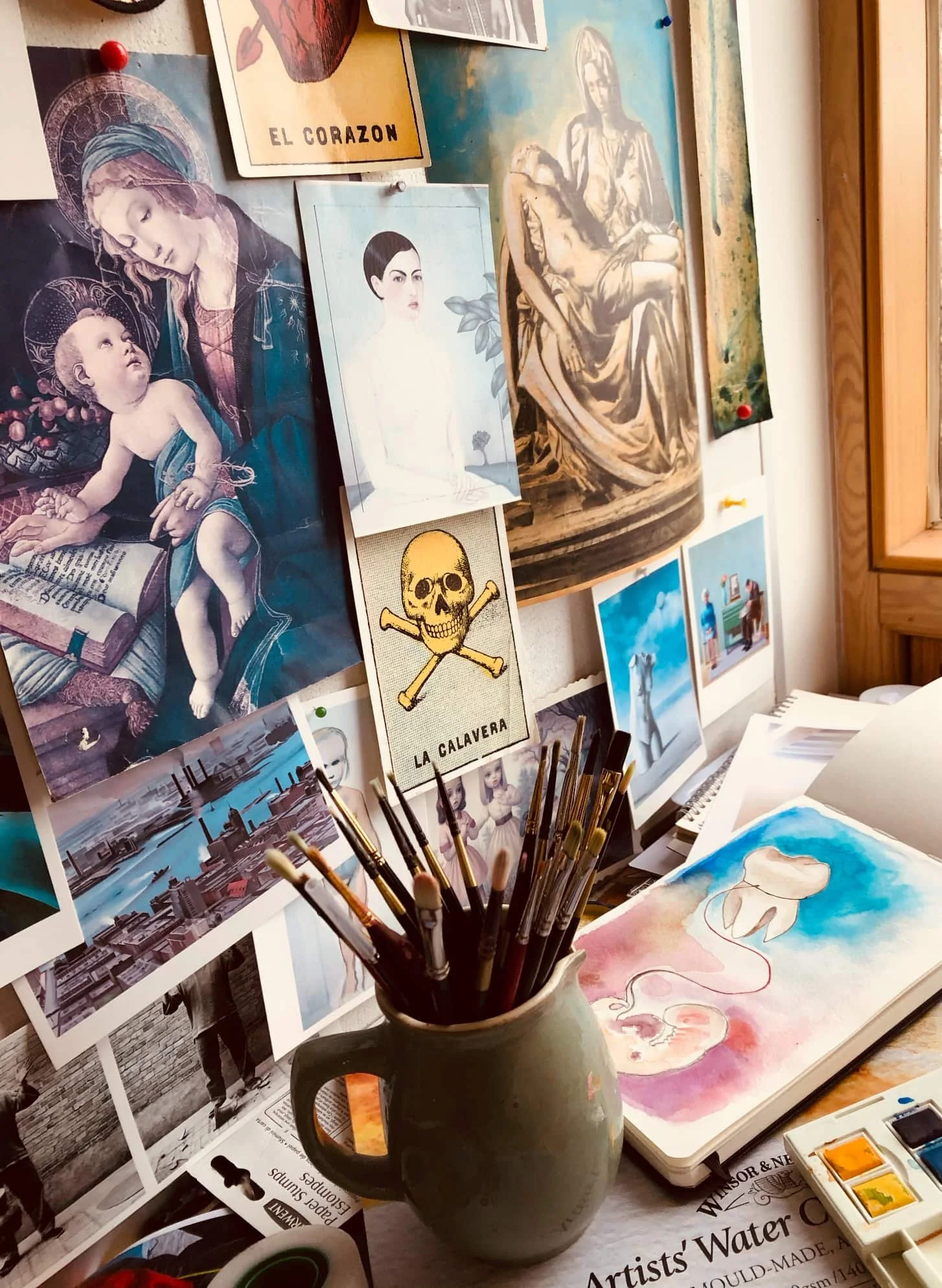 An artist's workspace with various paintings and drawings displayed on the wall. In the foreground, there is a container holding paintbrushes. On the table, there's an open watercolor set, a sketchbook with a watercolor painting of a tooth, and printed images of art and photography, including religious and skull artwork.