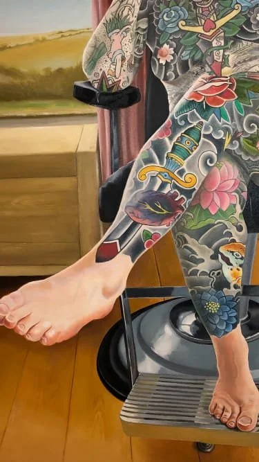 Tattooed arm and leg of a woman seated on a stool, showcasing colorful traditional Japanese style tattoos including roses, a dagger, a frog, waves.