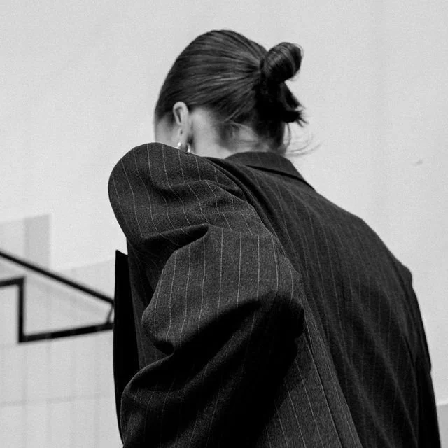 A woman with her hair tied in a bun, wearing a pinstripe blazer, facing away from the camera with her head slightly down.