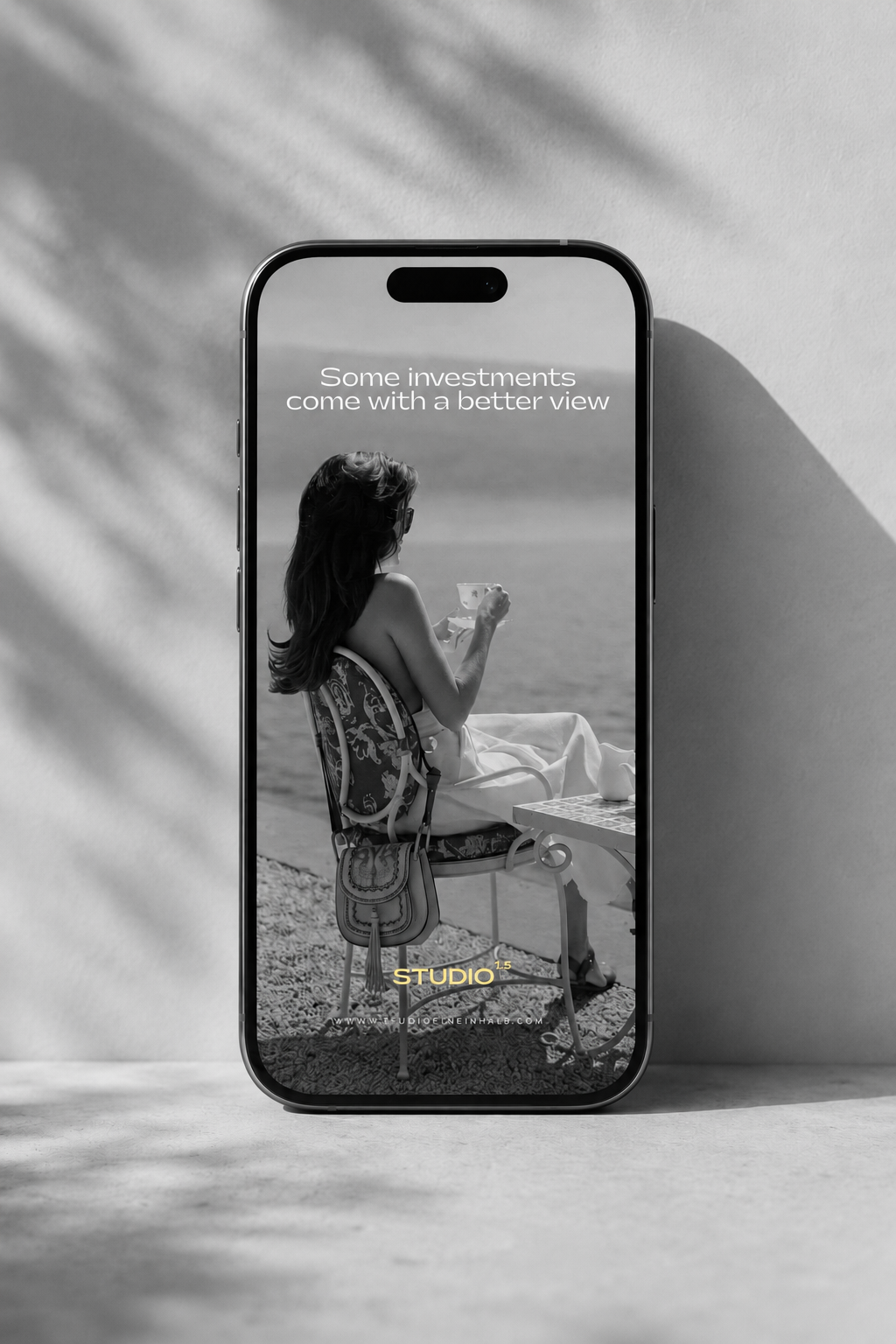 A smartphone displaying a black-and-white photo of a woman in sunglasses, sitting at a seaside table with a drink, with the text 'Some investments come with a better view' overlaid.