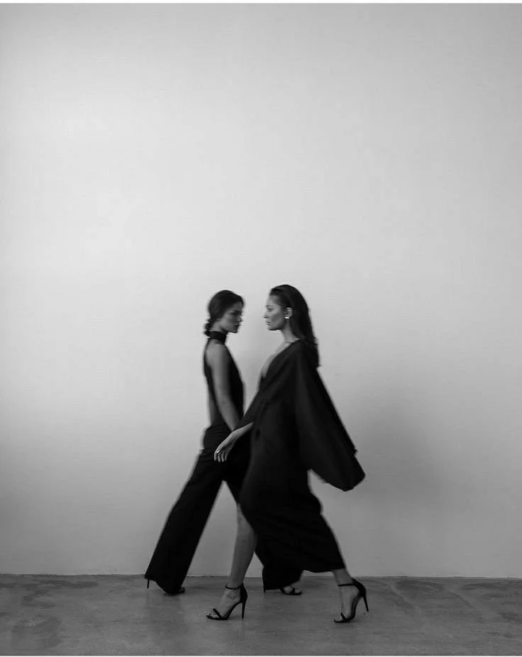 Two women walking together in black clothing and high heels, with one wearing a long coat, against a plain wall.