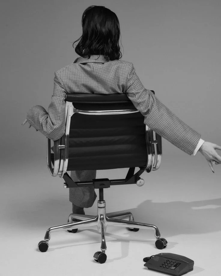 Rear view of a woman sitting in an office chair, wearing a checkered blazer, with a phone on the floor in front of her, reaching out with one arm.