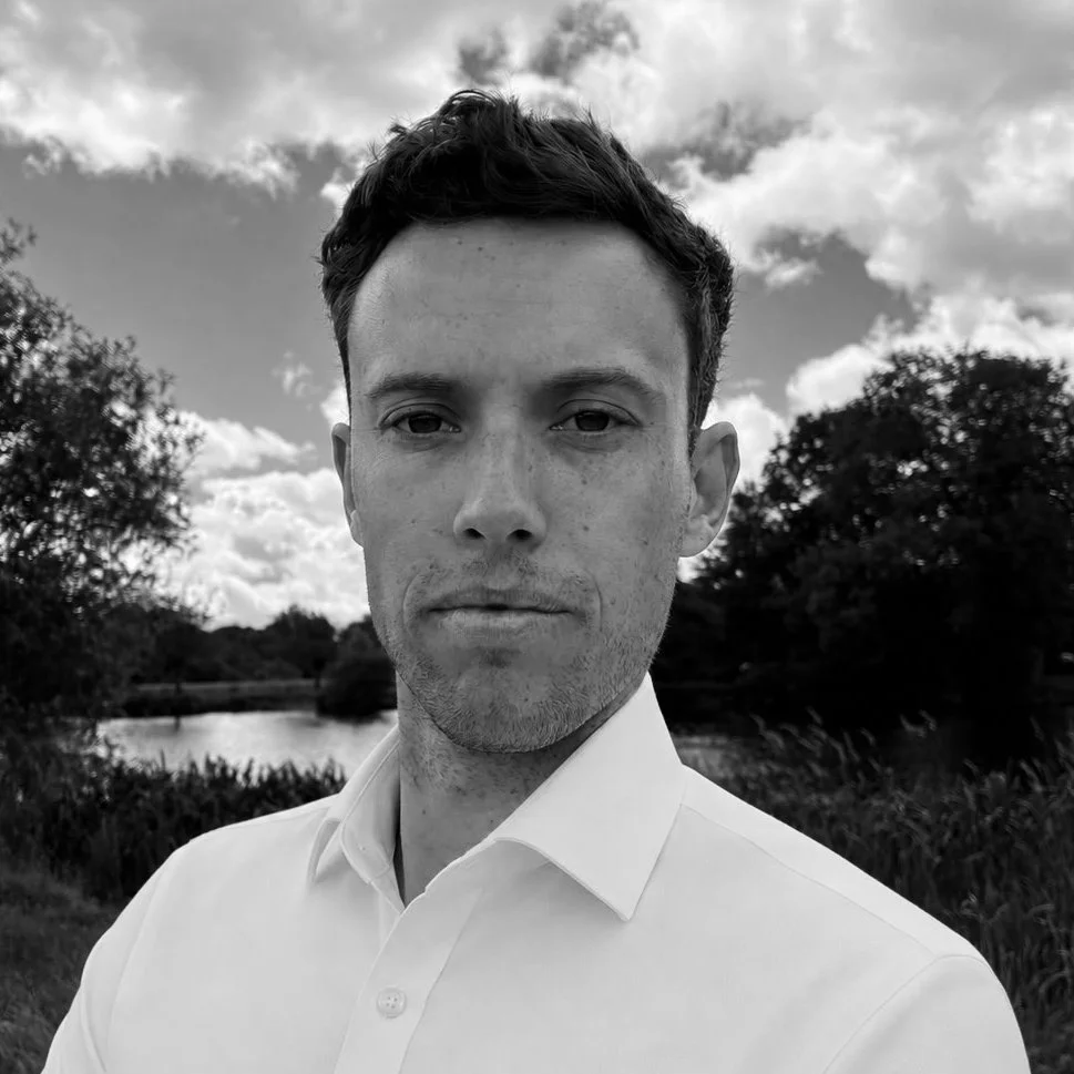A black and white photo of a young man with short dark hair, wearing a white dress shirt, standing outdoors near a body of water with trees and partly cloudy sky in the background.