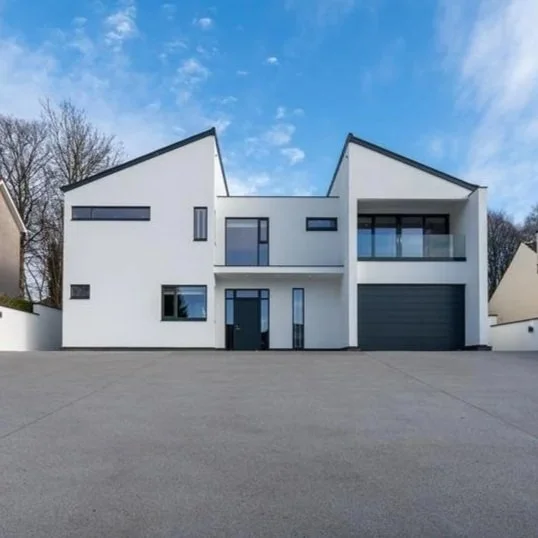 Modern white house with angular rooflines, large windows, and a driveway.