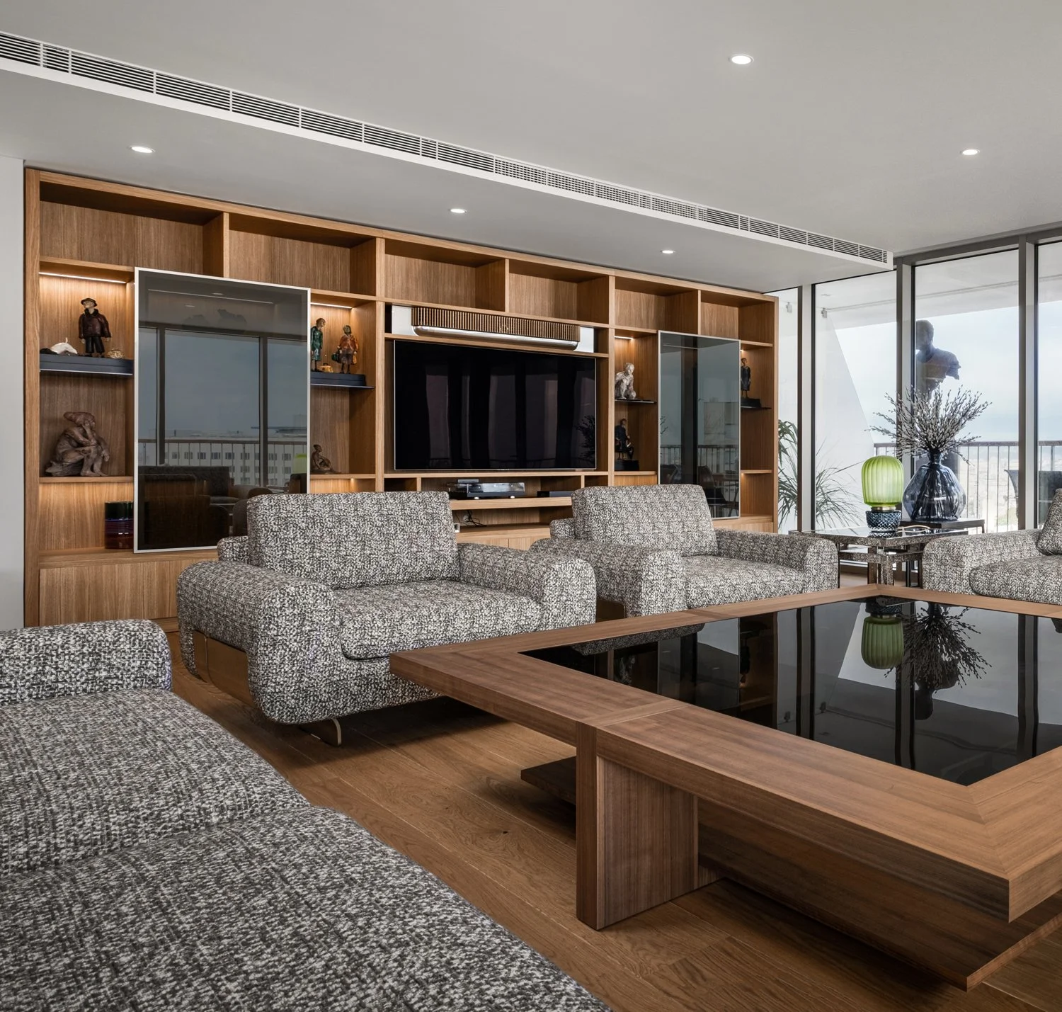Modern living room with patterned sofas, a wooden coffee table with a glass top, a wall-mounted flat-screen TV, built-in wooden shelves with decorative figurines, and floor-to-ceiling windows with a balcony view of the city.