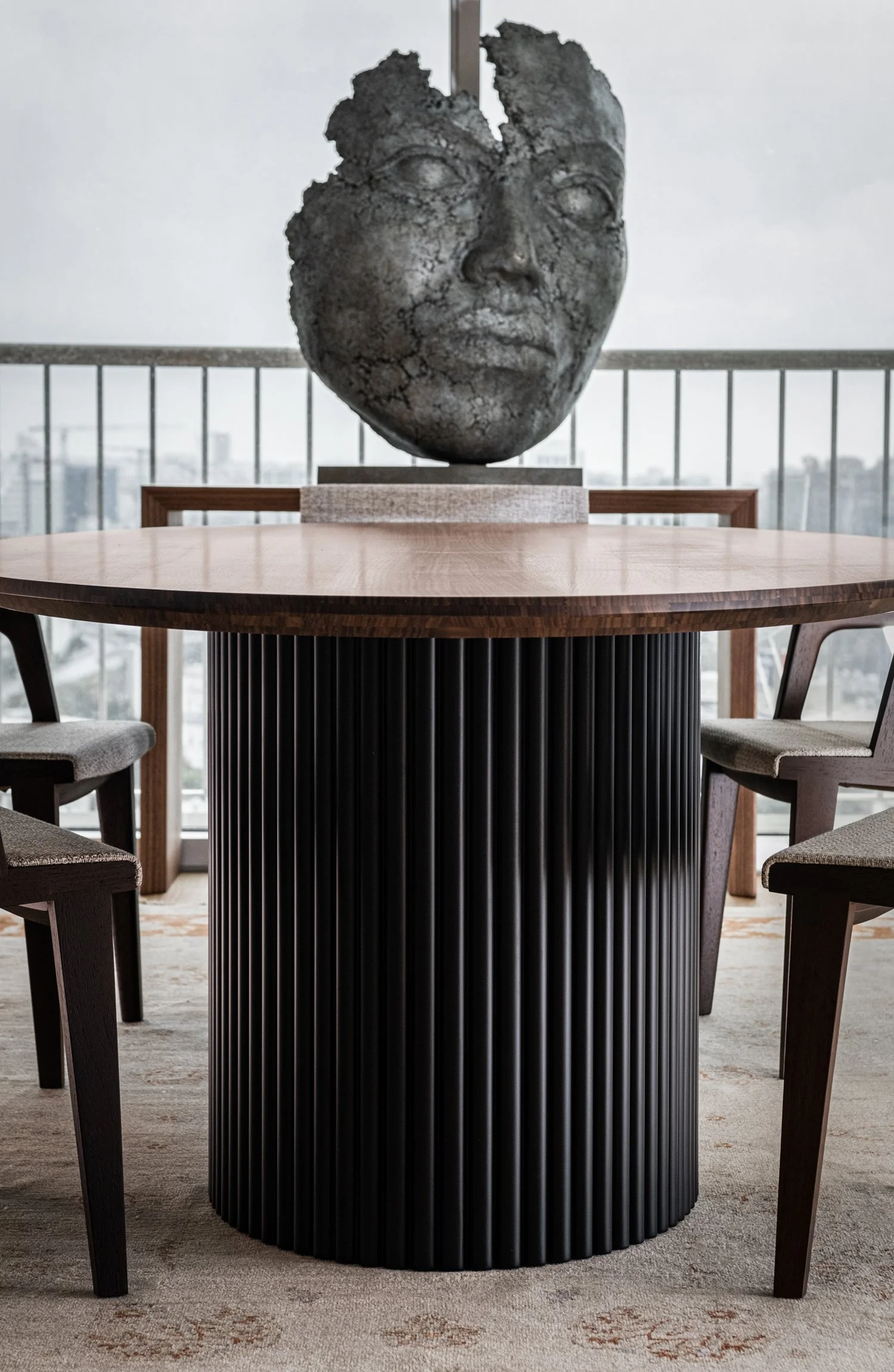 Modern round wooden dining table with a black ribbed cylindrical base, surrounded by four wooden chairs with upholstered seats, on a beige patterned carpet. In the background, a large sculpture of a talking face with cracked texture and closed eyes, 