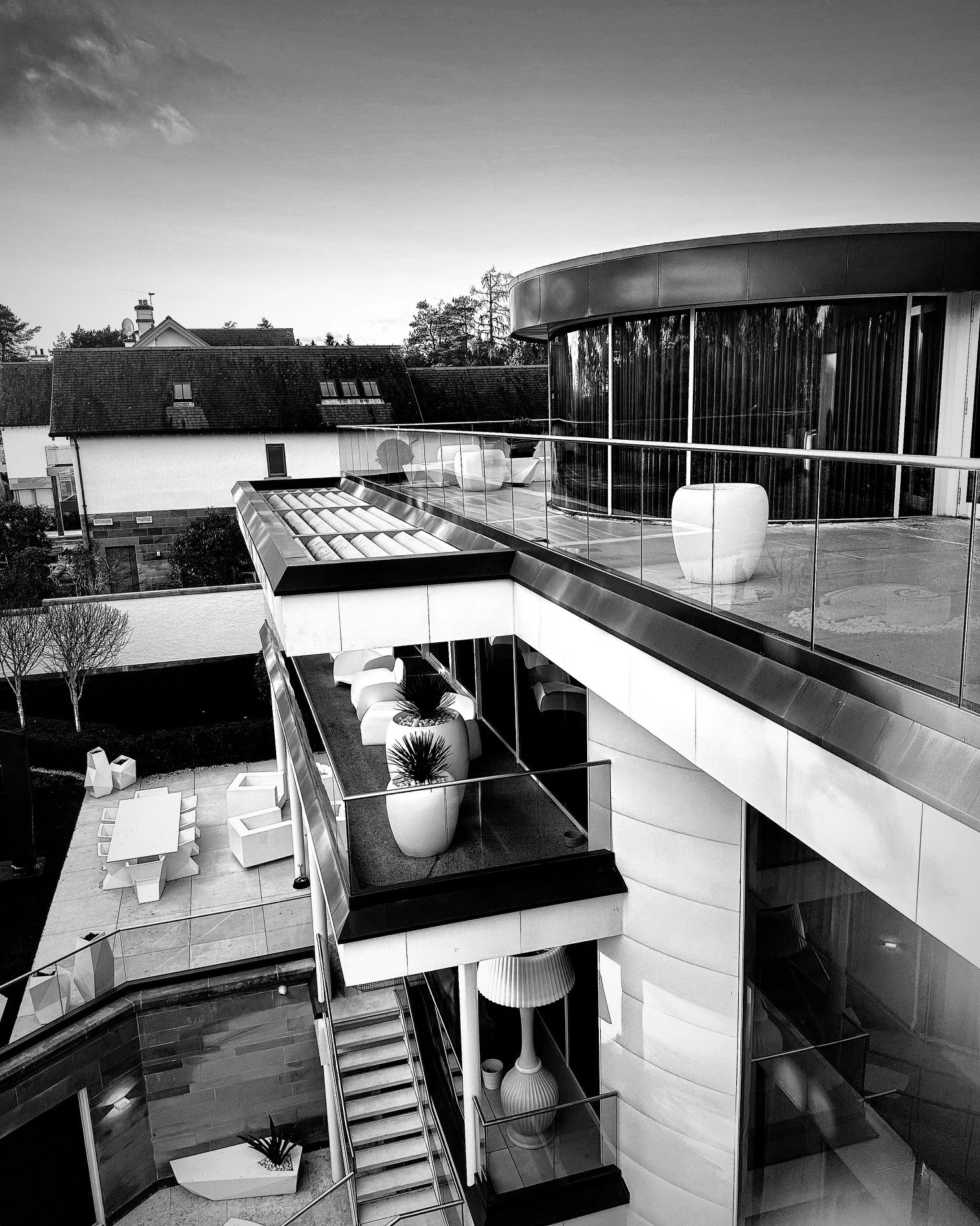 Modern multi-story house with outdoor balcony, potted plants, glass railings, and outdoor seating area with white chairs and tables, overlooking neighboring rooftops.