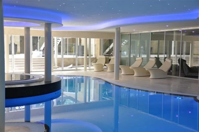 Indoor swimming pool area with lounge chairs and glass walls, illuminated with blue and white lighting.