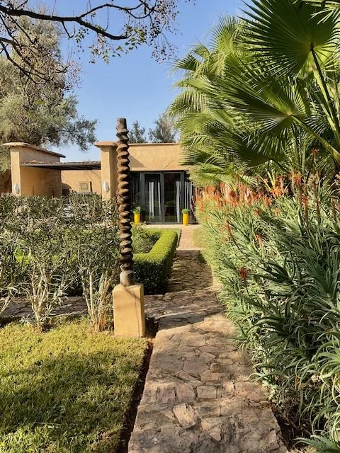 Stone pathway leading to a house with large windows, surrounded by lush landscaping including palm trees, bushes, and flowers.