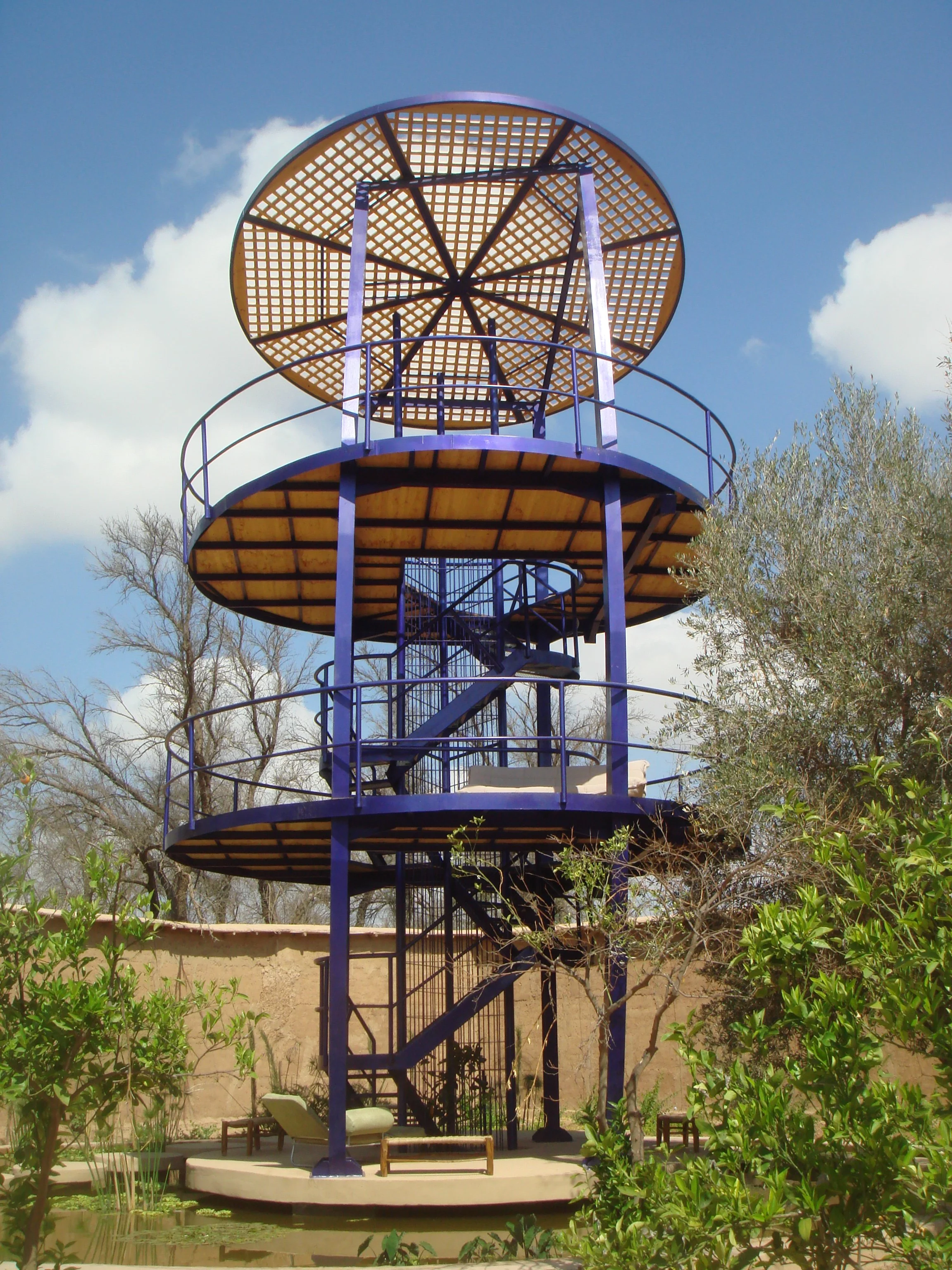 A multi-level blue and yellow tower structure with a spiral staircase, an umbrella-shaped top, and surrounding greenery