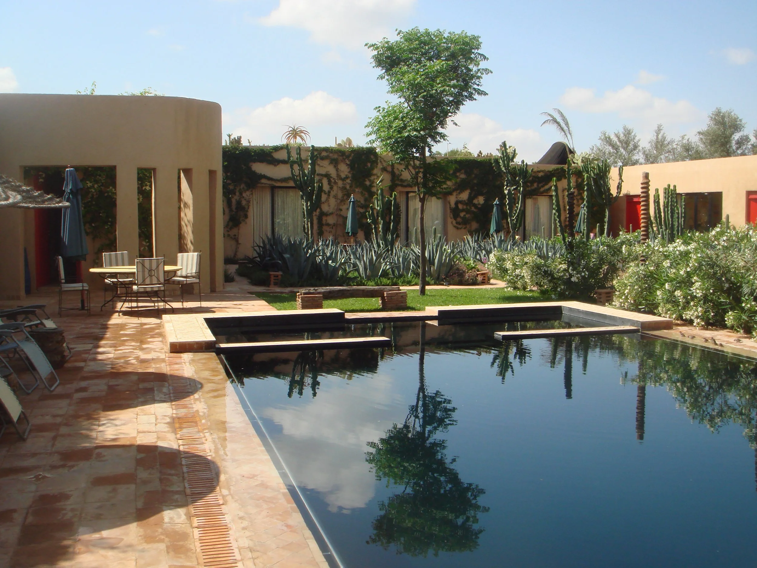 A courtyard with a swimming pool in Morocco designed by Julian Hunter Architects, patio furniture, green plants, and a beige building with rounded walls and rectangular windows, under a partly cloudy sky.