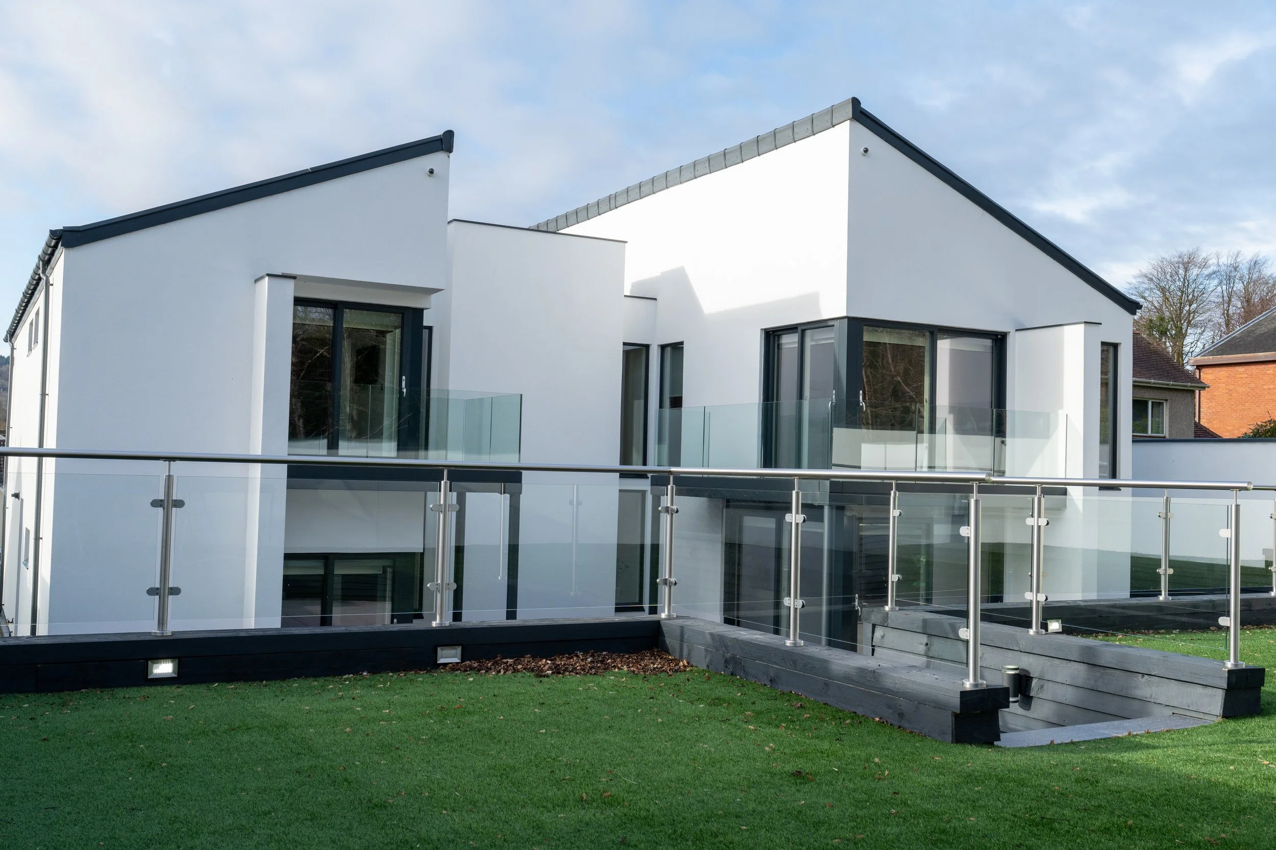 Modern white residential building with large glass windows and glass balcony railings, surrounded by a green lawn, under a partly cloudy sky.
