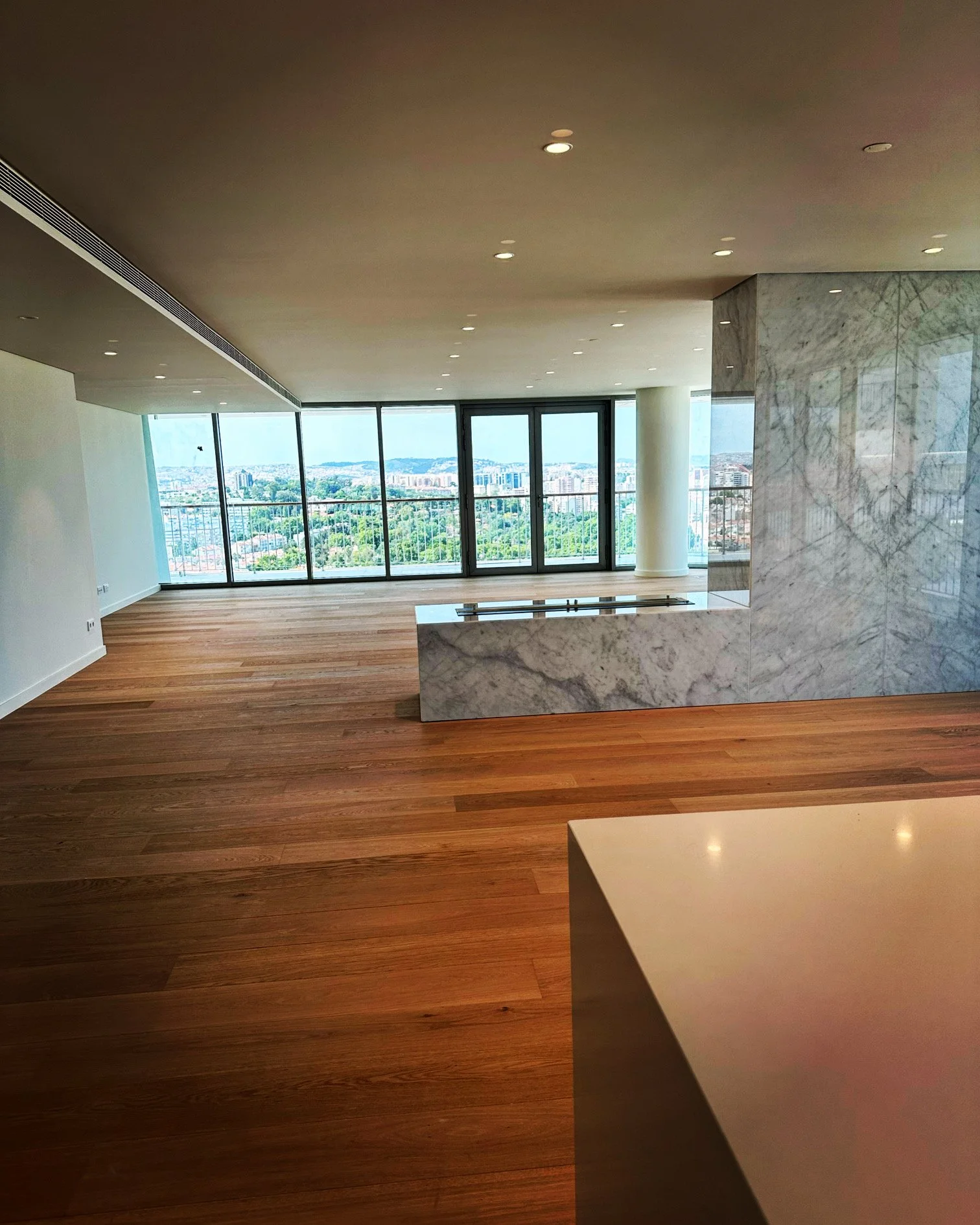 Interior of a modern high-rise apartment in Lisbon with large windows overlooking a cityscape with greenery, wooden flooring, recessed ceiling lights, and marble accents.