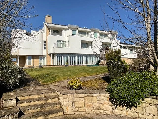 Modern white apartment building with multiple balconies and large windows, surrounded by a landscaped yard with stairs, shrubs, and a children's slide, under a clear blue sky.