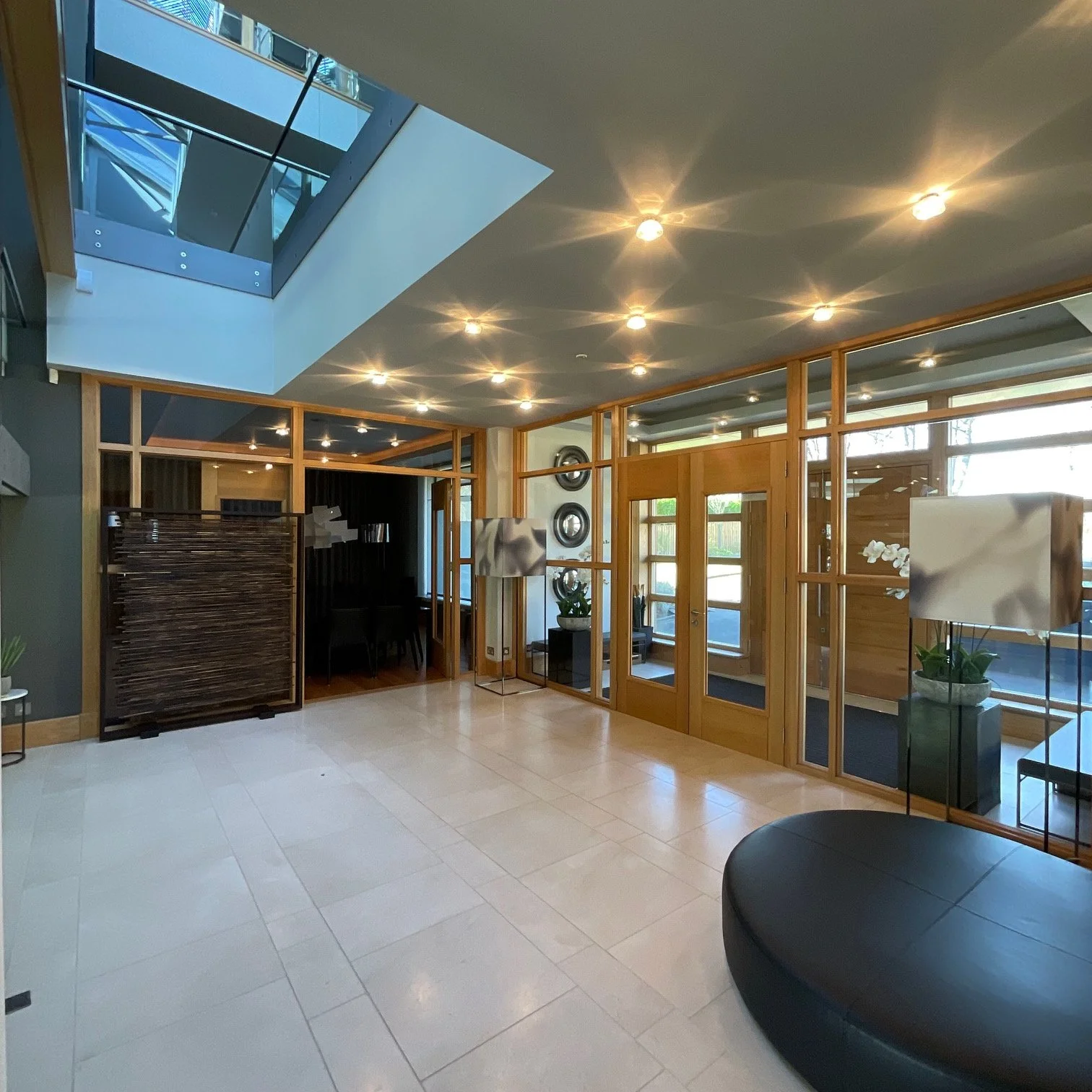 Interior of a modern building lobby with hanging lights, wooden and glass partitions, indoor plants, and a black leather rounded seating area.