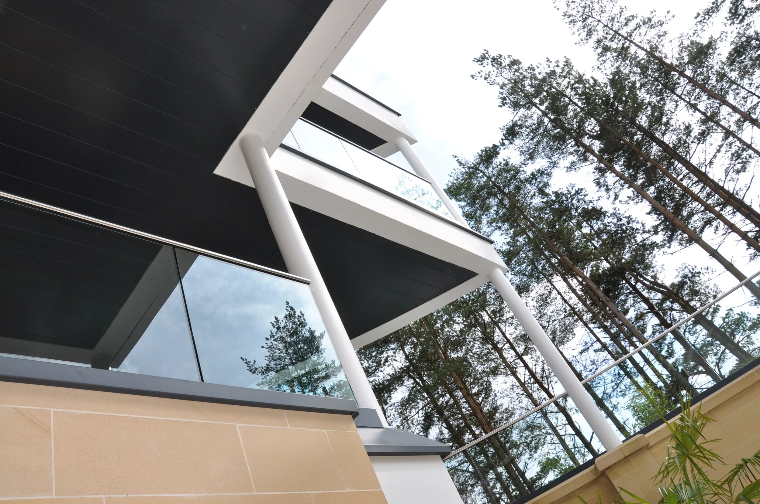 Modern building with large windows, white balconies, and black panels, surrounded by tall trees, under a partly cloudy sky.