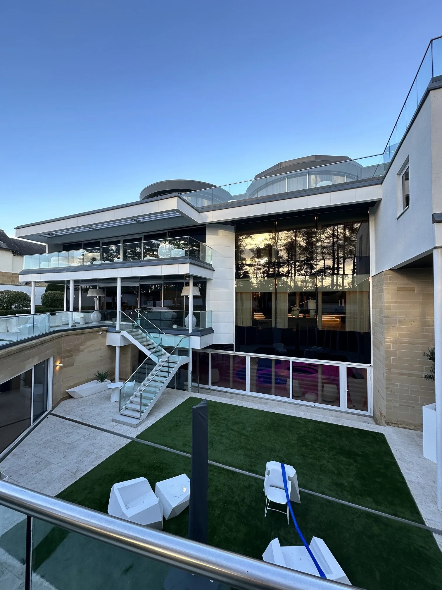 Modern house with glass balconies, large windows, and a patio area with chairs and artificial grass