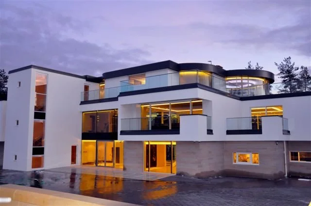 Large modern house with large glass windows and balconies, illuminated from within, during dusk with a wet pavement reflecting the lights.