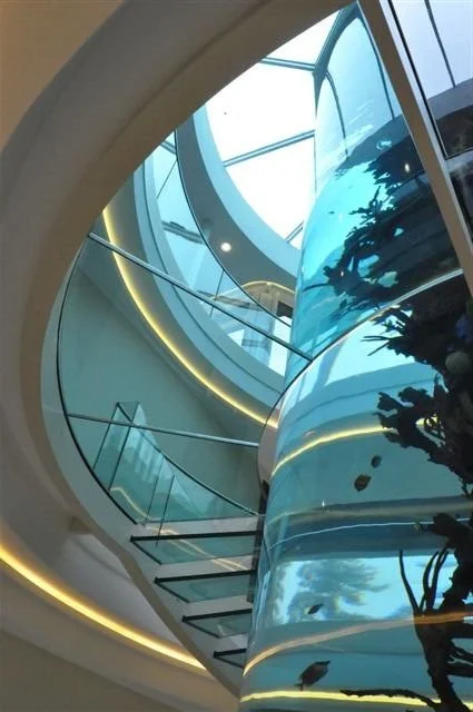 Interior view of a modern spiral staircase with glass railings and a circular glass aquarium, illuminated with LED lighting.