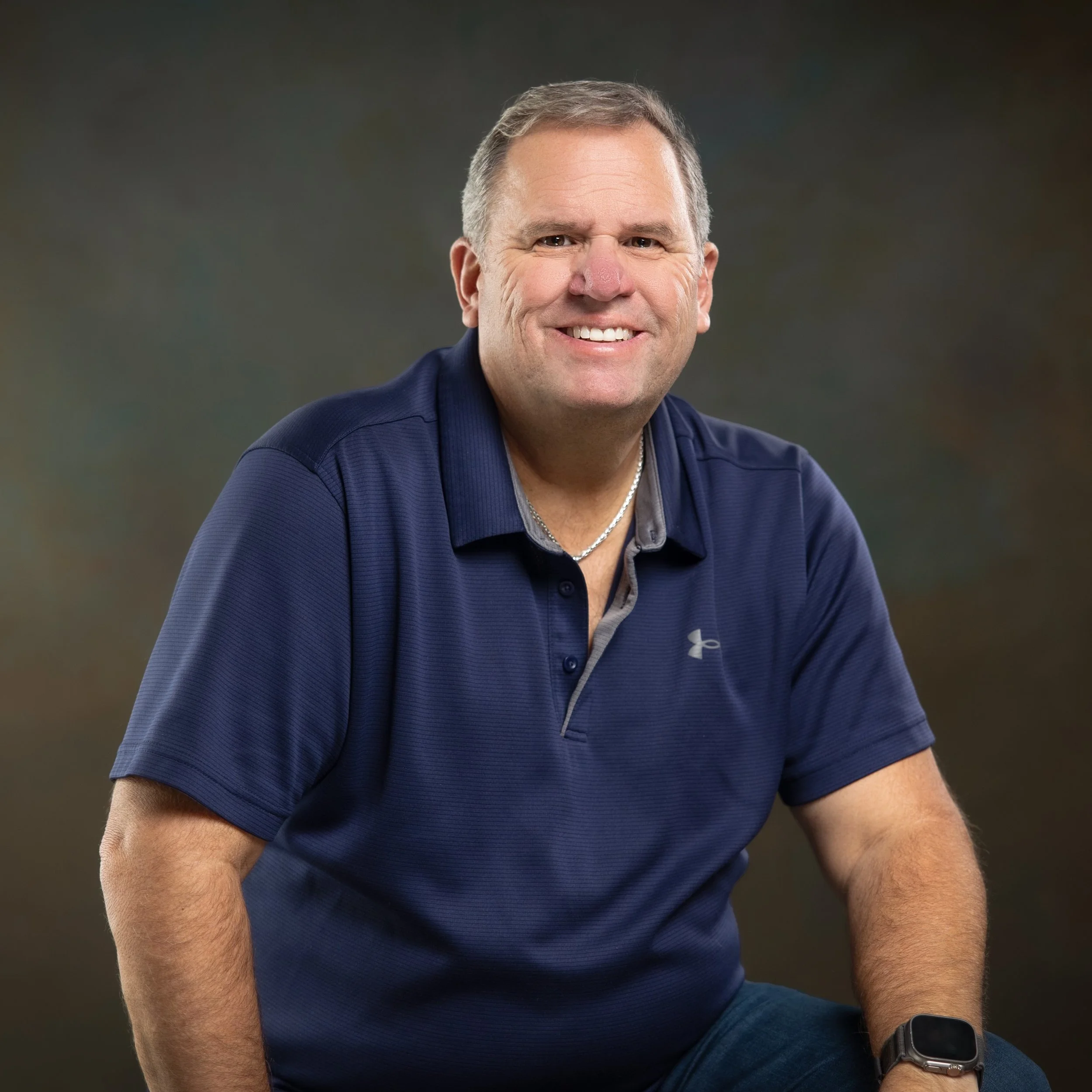 A man with short brown hair smiling and wearing a navy blue polo shirt with a light gray collar, sitting against a dark blurred background.