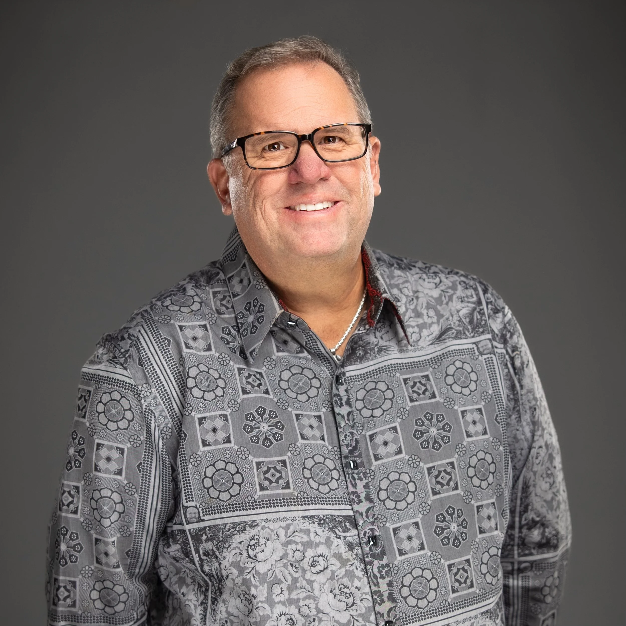 A smiling middle-aged man with light skin, short gray hair, and black rectangular glasses. He is wearing a patterned button-up shirt with floral and geometric designs in shades of gray and black, and a silver chain necklace. The background is plain dark gray.