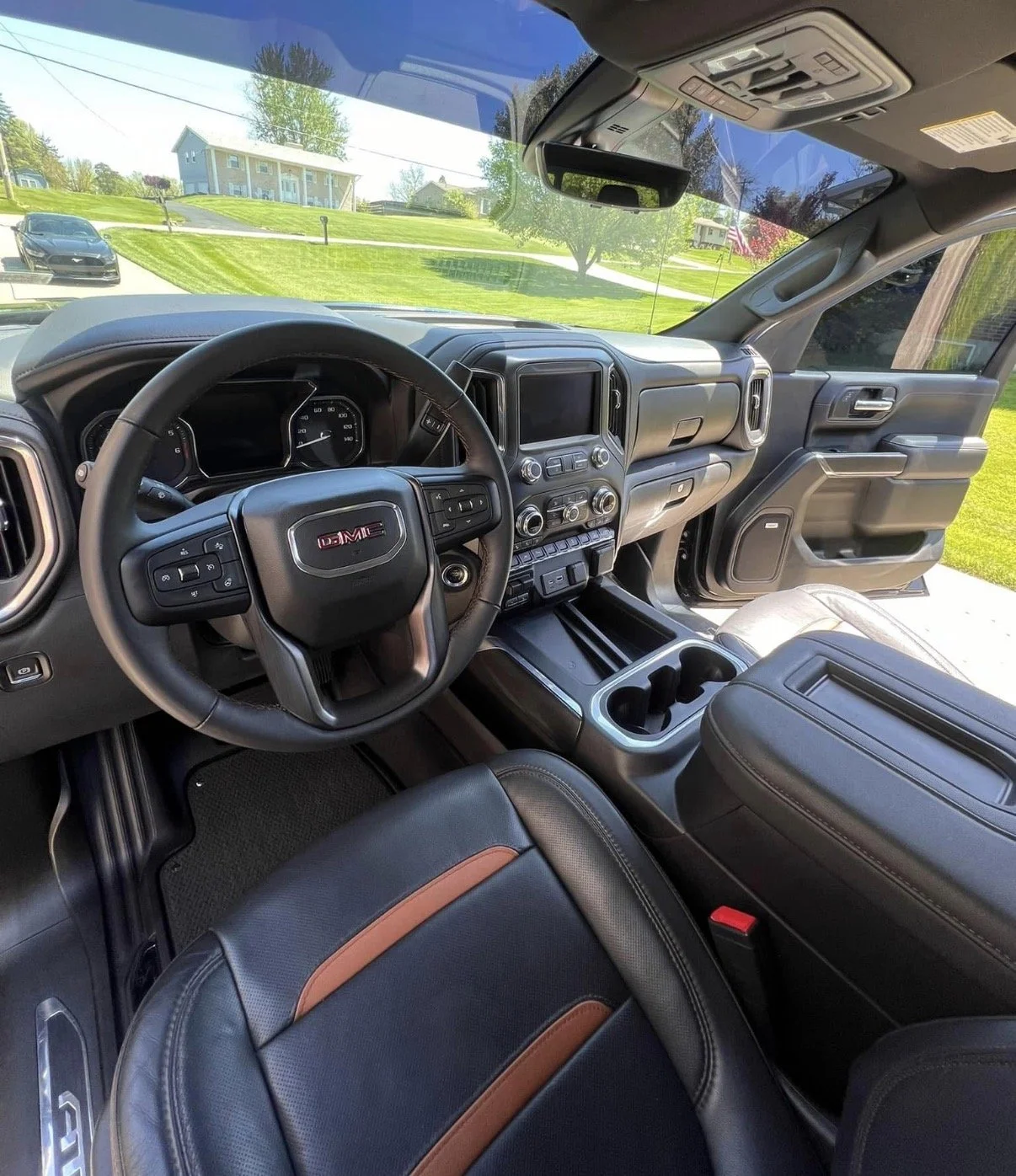 Interior view of a GMC vehicle with a black steering wheel, dashboard, and center console, parked in a suburban neighborhood with houses and green lawns visible through the windshield.