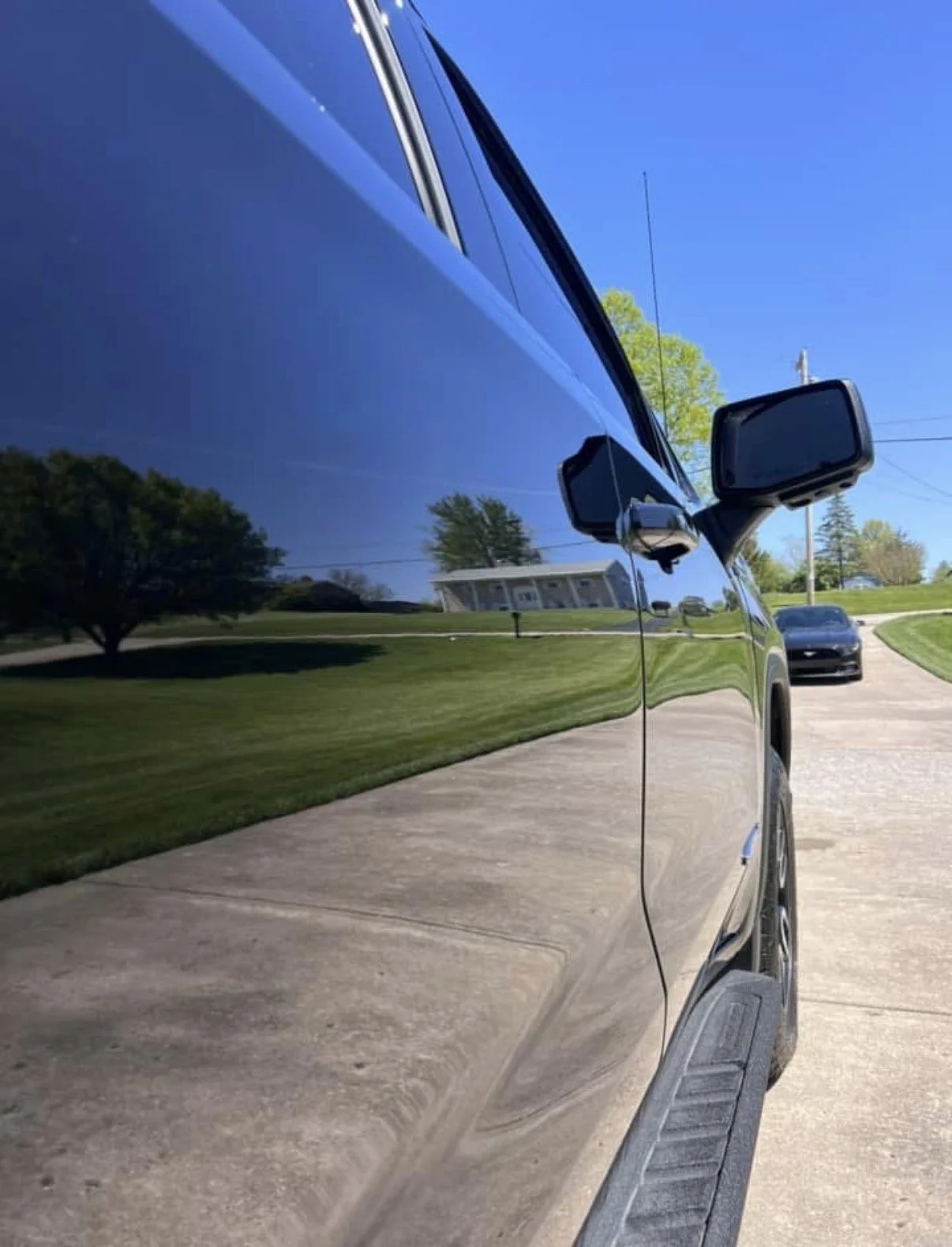 A close-up side view of a black vehicle with a highly reflective surface, showing reflections of green lawns, a tree, a house, and a blue sky.
