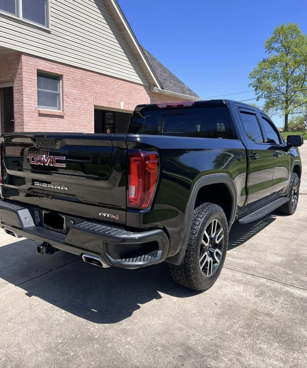 A black GMC Sierra AT4 pickup truck parked in a driveway next to a house with a beige and pink brick exterior, under a clear blue sky and a green tree.