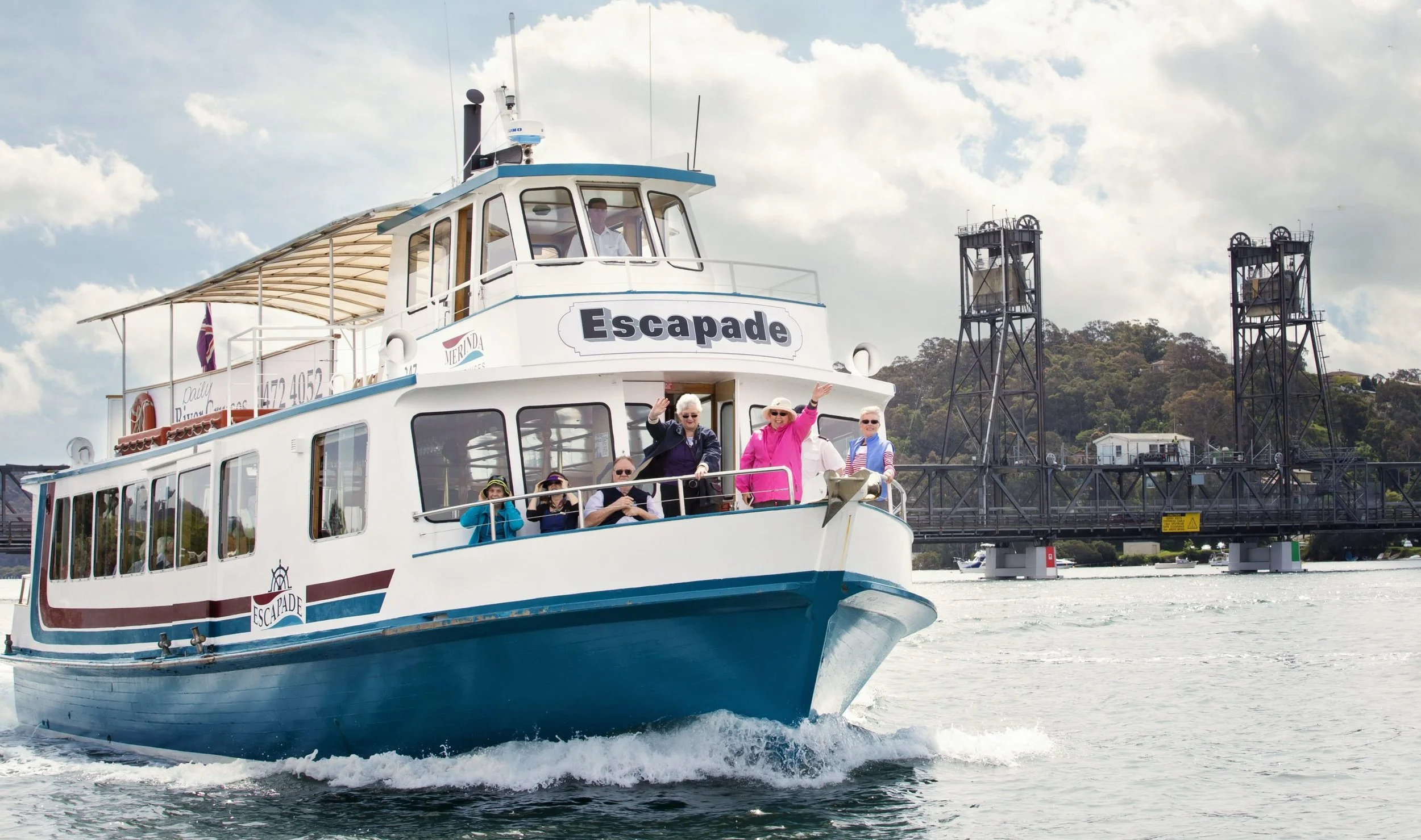 A group of people on a white and blue boat named Escapade sailing on the water with a bridge and hill in the background, under cloudy sky.