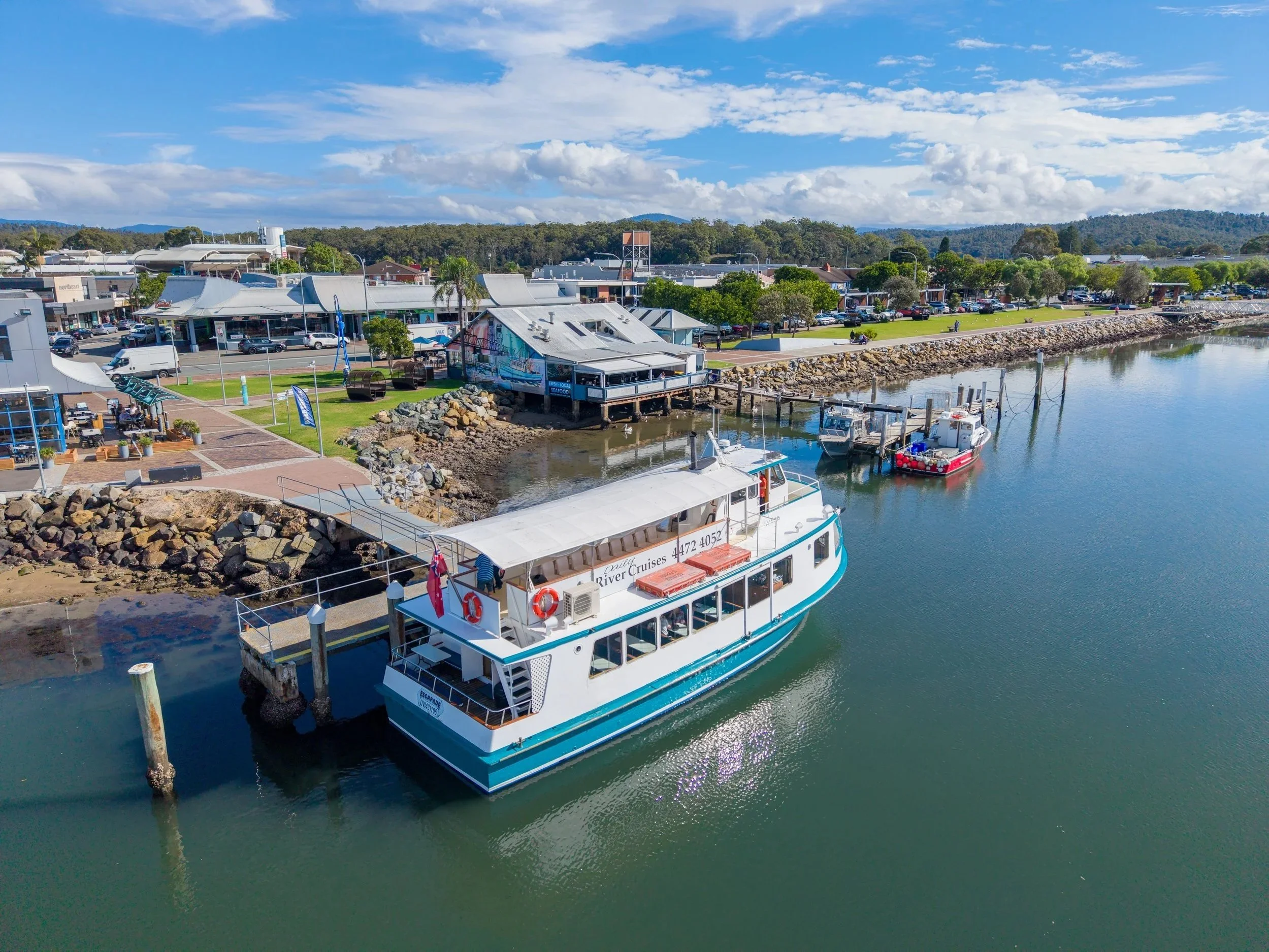 A waterfront scene with boats docked along a marina and a shopping area with cars parked, surrounded by trees and hills under a blue sky with clouds.