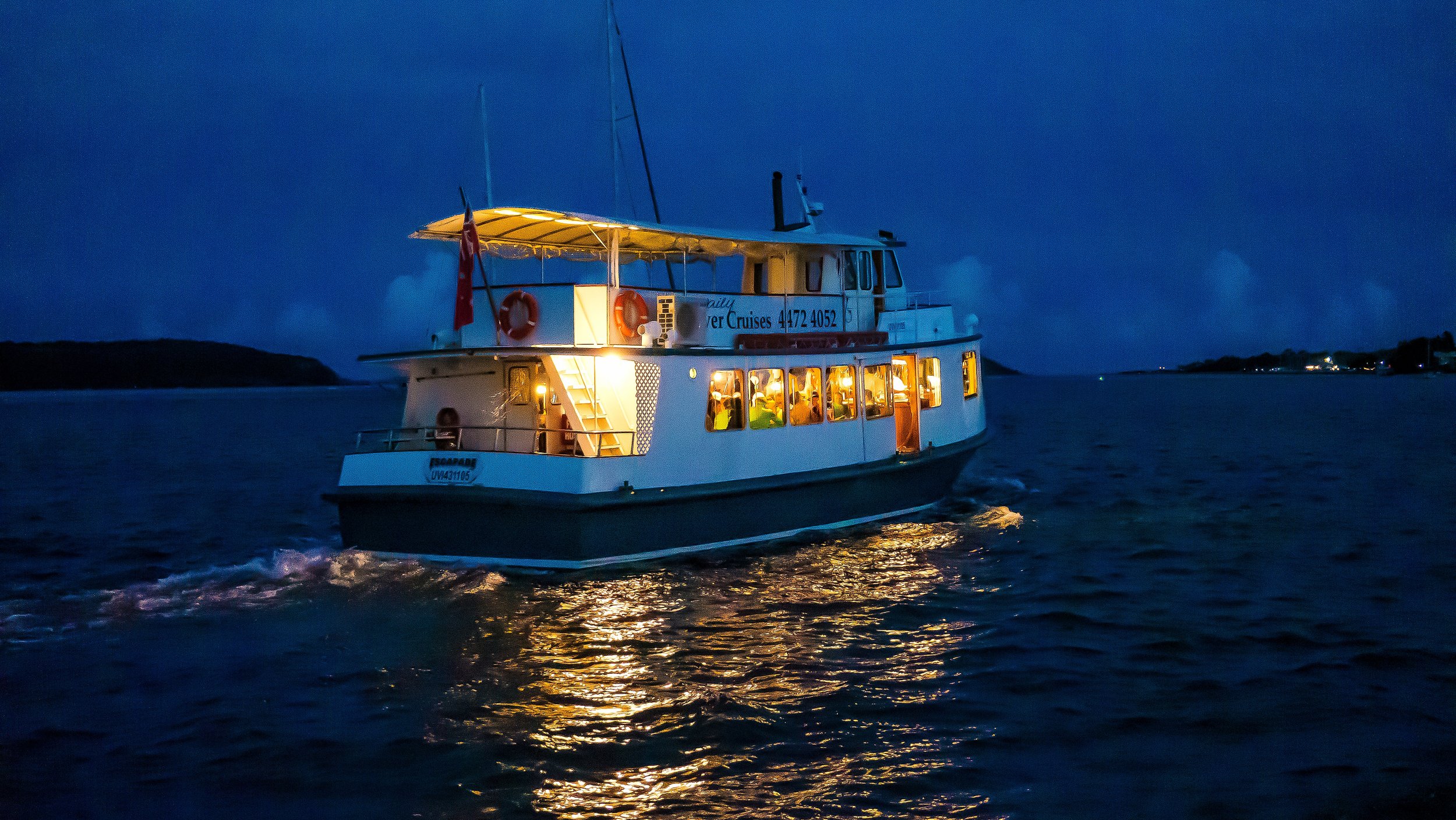 A boat with yellow lights on deck cruising on the water during the night, with dark sky and distant shoreline visible.